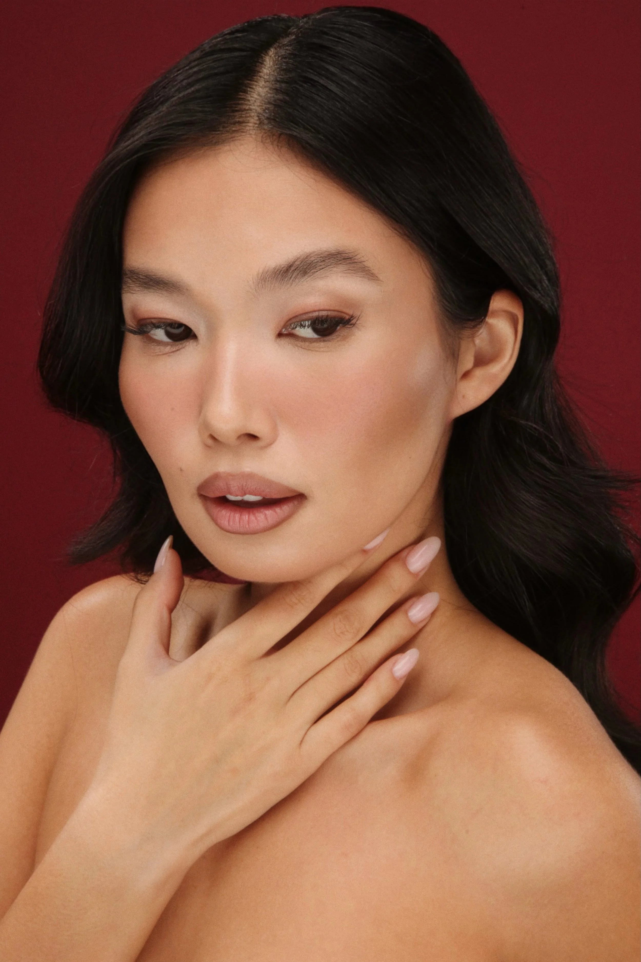 Ritratto di una donna con capelli neri, pelle luminosa, trucco naturale e mani poggiate sulla spalla, sfondo rosso.