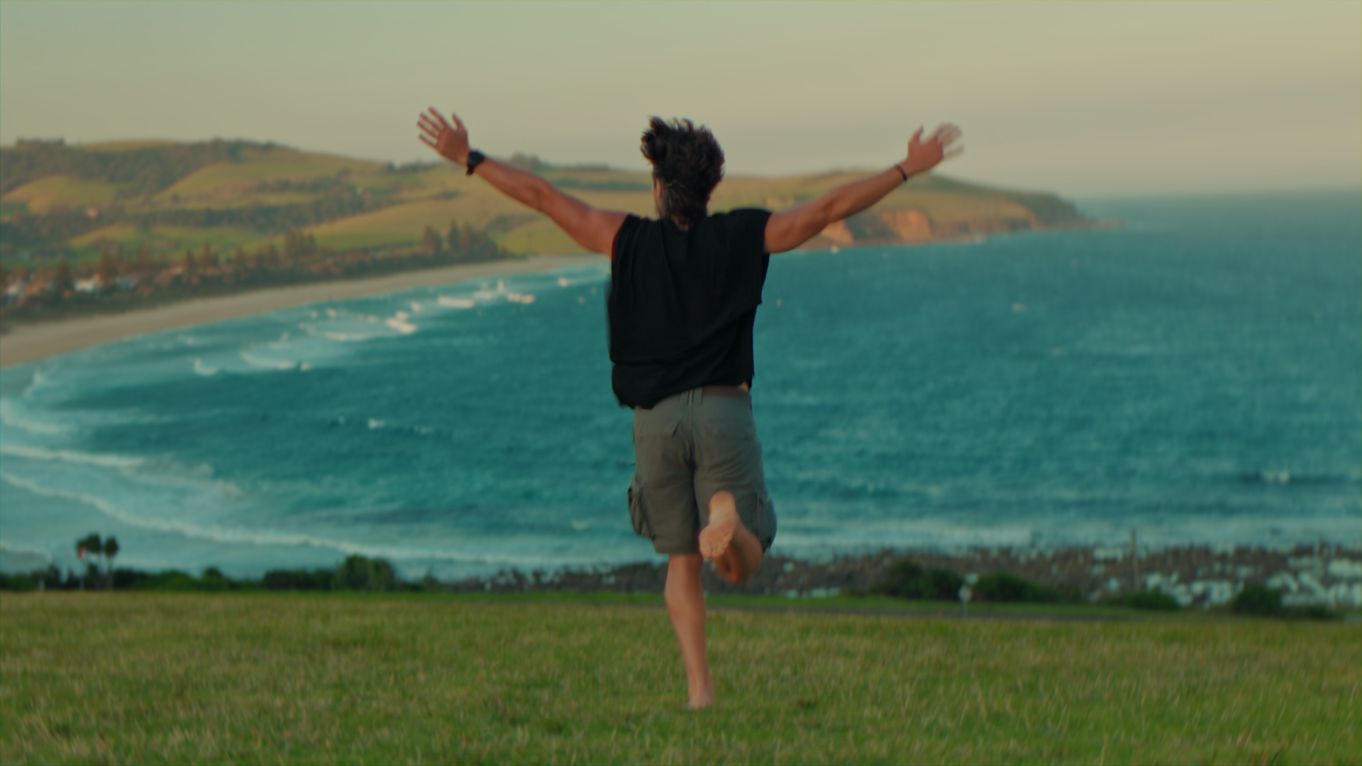 Un uomo con maglietta nera e pantaloncini corti si trova su un prato, con le braccia aperte e un piede sollevato, di spalle in primo piano, mentre guarda il mare e le colline sullo sfondo