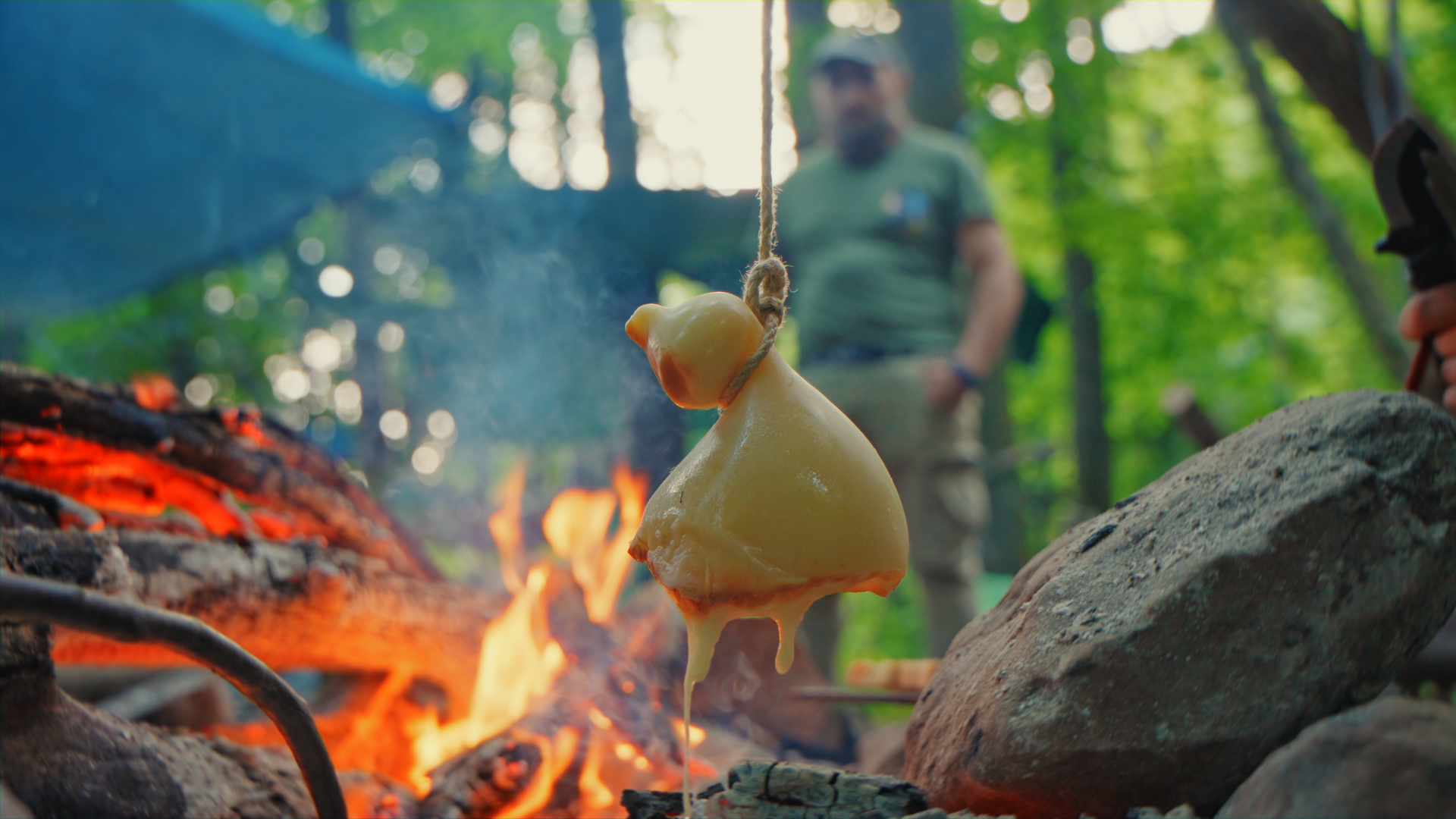 Formaggio fuso che cola su un caminetto acceso in un bosco, con persone sullo sfondo.