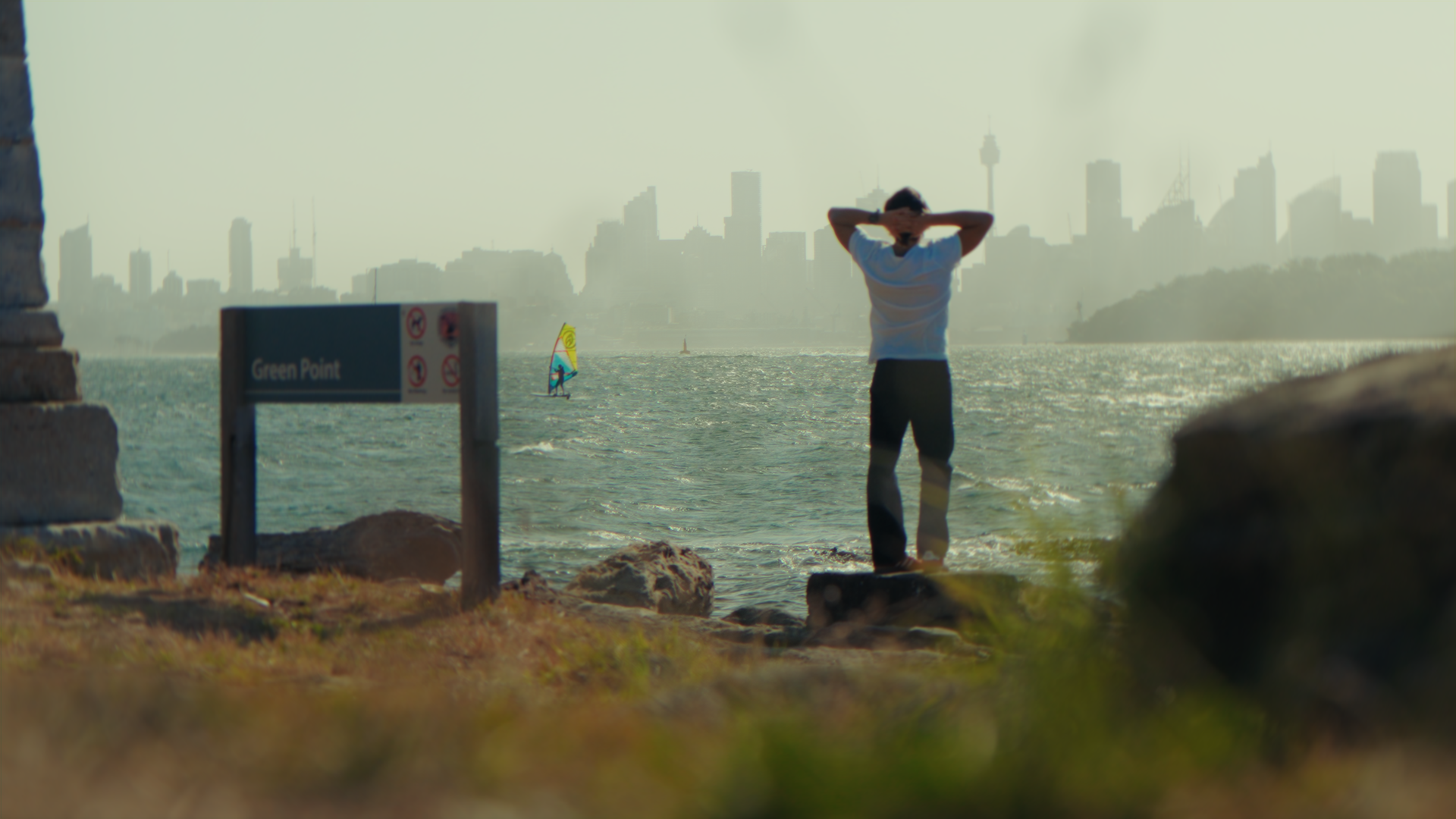 Una persona si trova sulla riva di un lago o mare, con alle spalle la silhouette di una città e un cielo filtrato dalla luce solare.