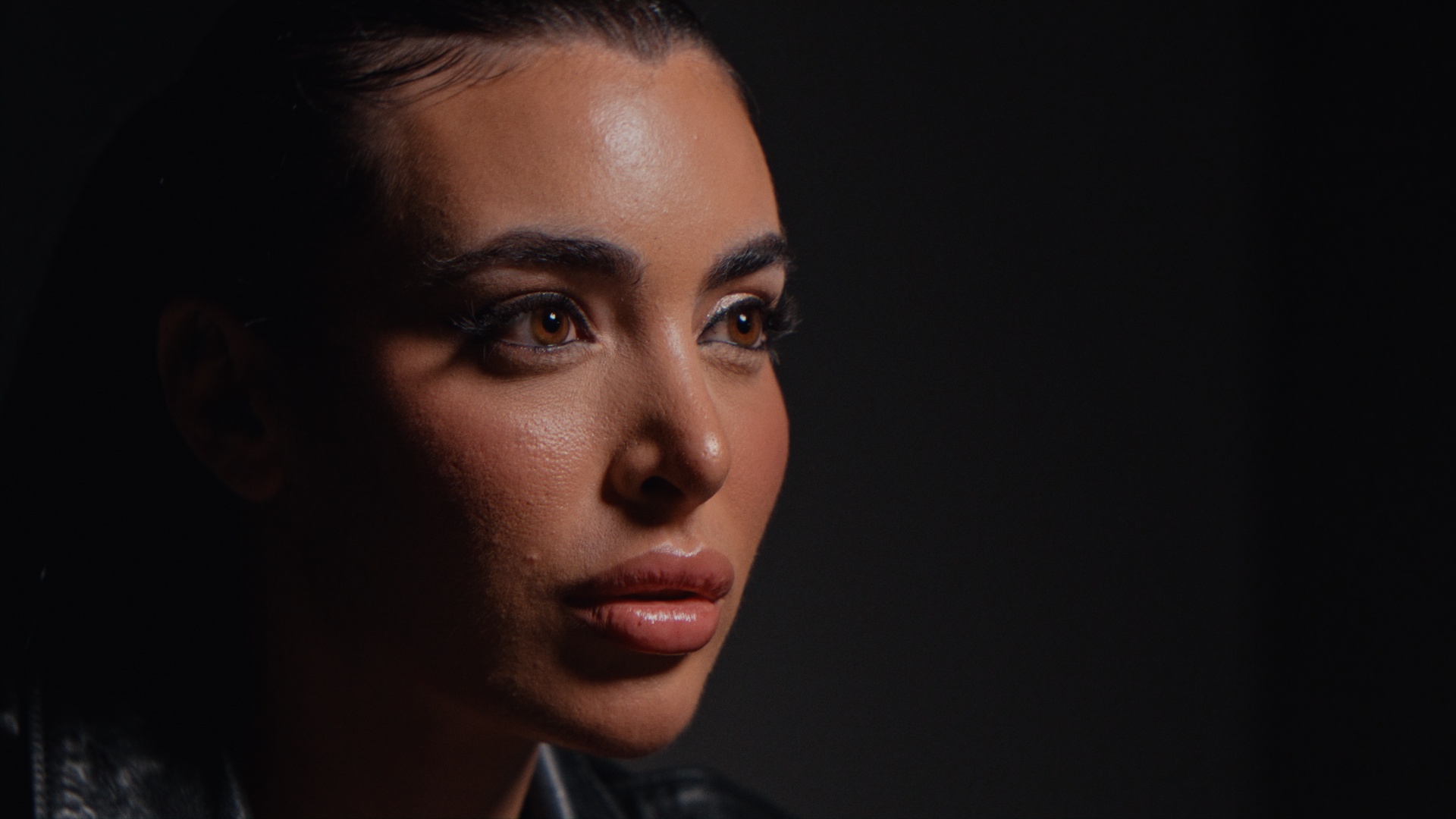 Primo piano di una donna con pelle olivastra, occhi marroni, trucco naturale e capelli scuri stretti all'indietro, su sfondo nero