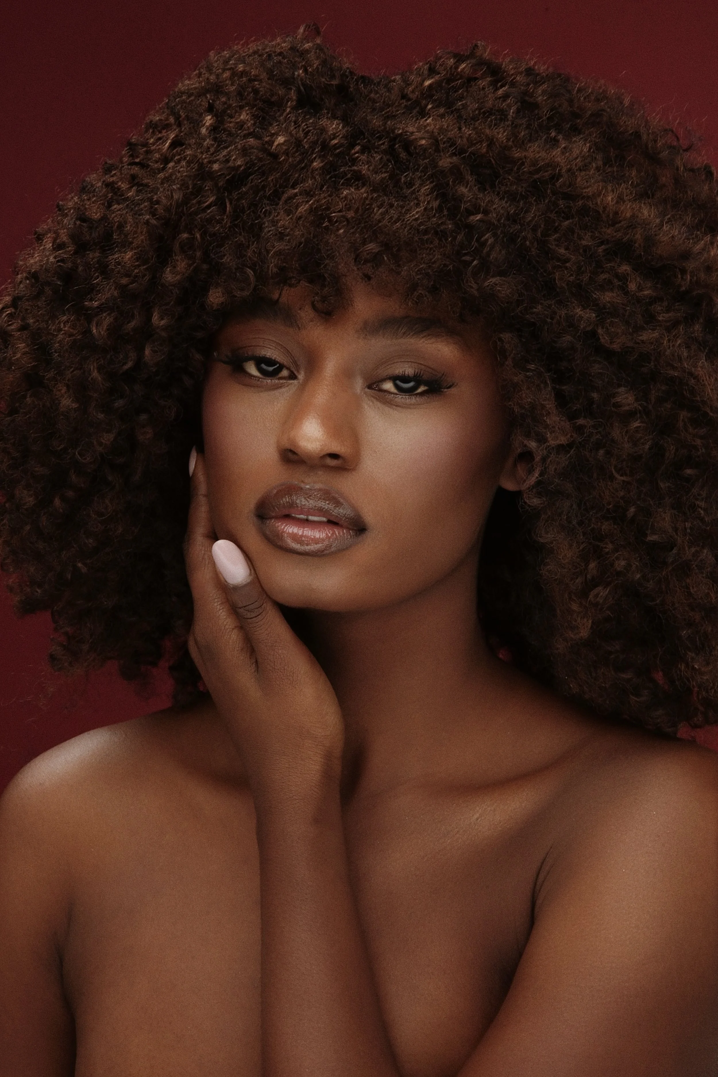 Ritratto di una donna con capelli ricci voluminosi, pelle liscia e truccata, che tocca delicatamente il viso con una mano, sfondo rosso scuro.
