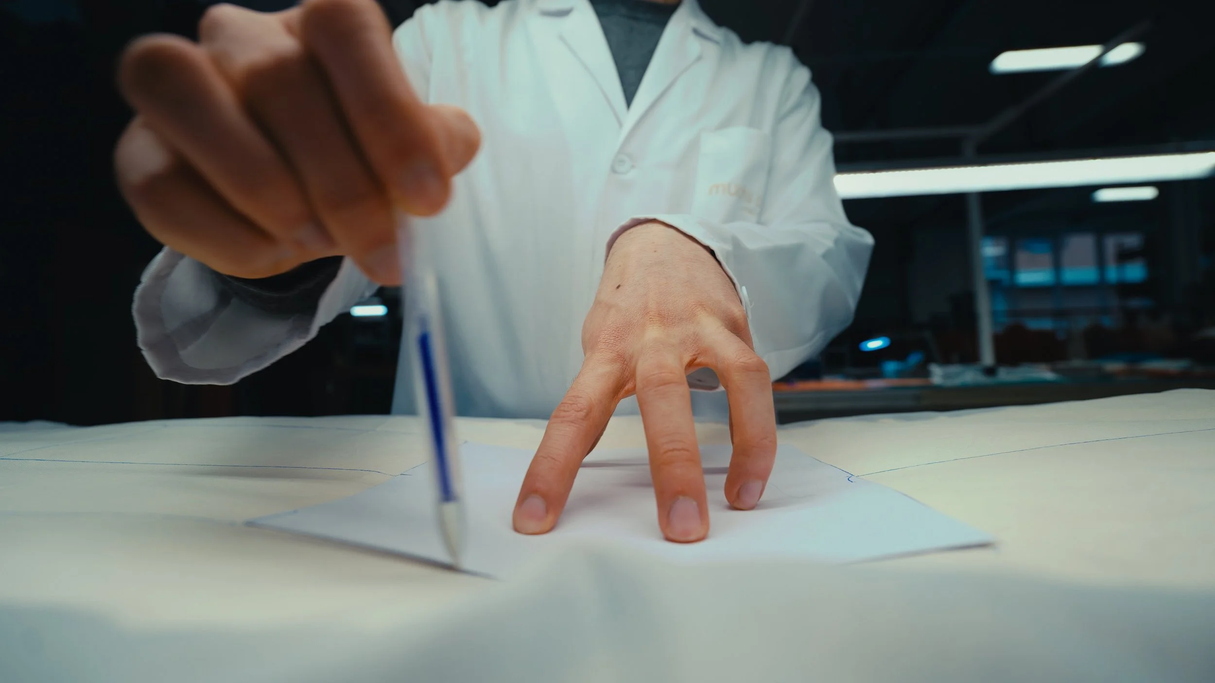 Una persona che disegna su un grande foglio di carta con un penna, in un ambiente di lavoro o laboratorio, indossando un camice bianco.