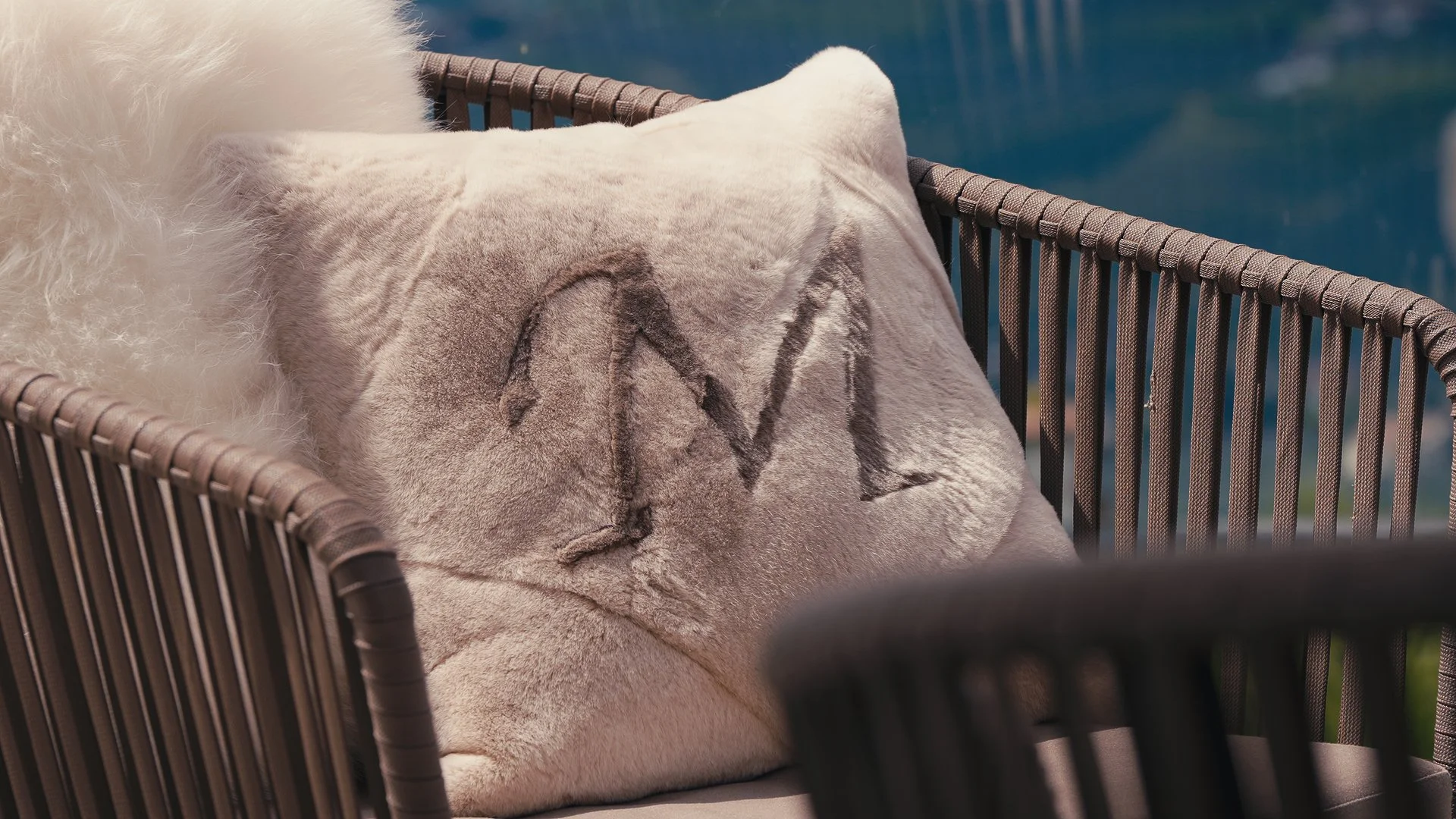 Cuscino di pelliccia beige con la parola 'LOVE' ricamata, posato su una sedia di vimini, con sfondo di acqua blu.