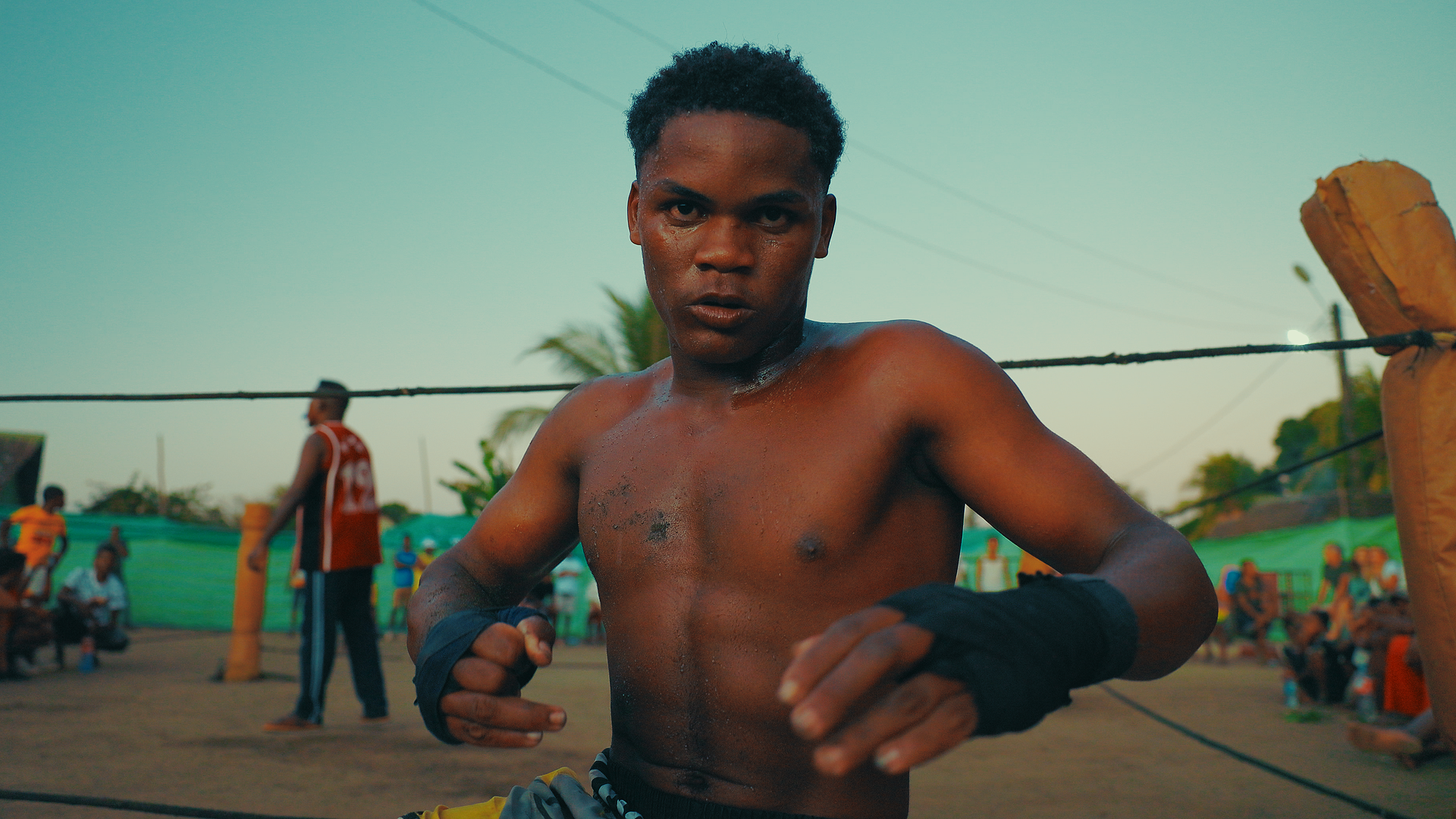 Un ragazzo magro e muscoloso, senza maglietta e con le mani avvolte in fasce nere, si trova sul ring di un'arena di boxe all'aperto, con uno sguardo deciso, in primo piano, mentre sullo sfondo ci sono altre persone sedute e in piedi, alcune di fronte a cabine o spazi delimitati, in un ambiente all'aperto con cielo azzurro e zone verdi.