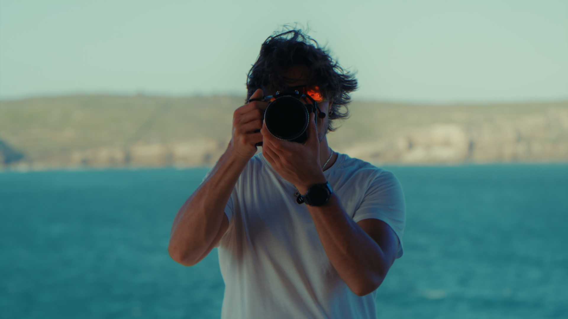 Un uomo con capelli scuri e ricci che indossa una maglietta bianca e un orologio nero sta scattando una fotografia con una fotocamera reflex vicino a un corpo d'acqua con una collina sullo sfondo.