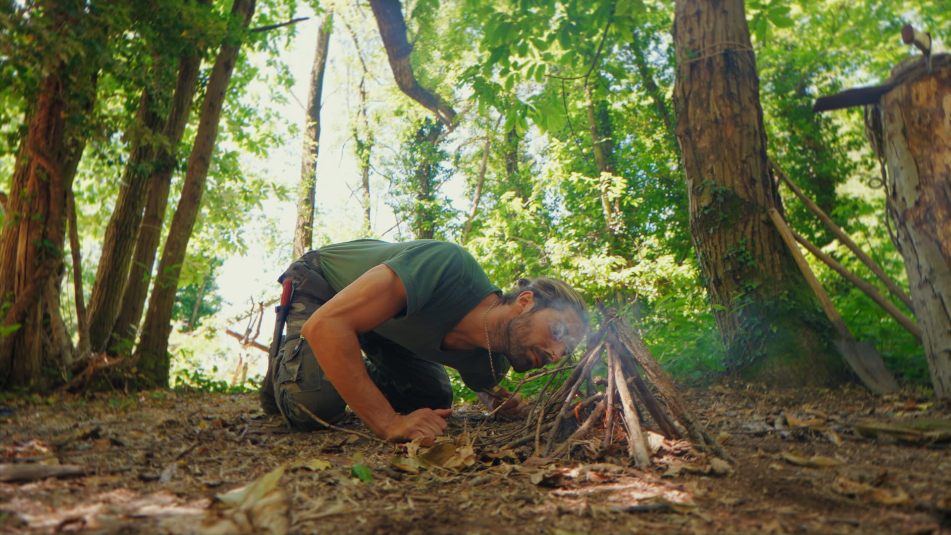 Uomo in un bosco che tende un falò con rami secchi. Alterna tra camicie verdi e pantaloni cargo. Attorno ci sono alberi alti e fogliame verde.