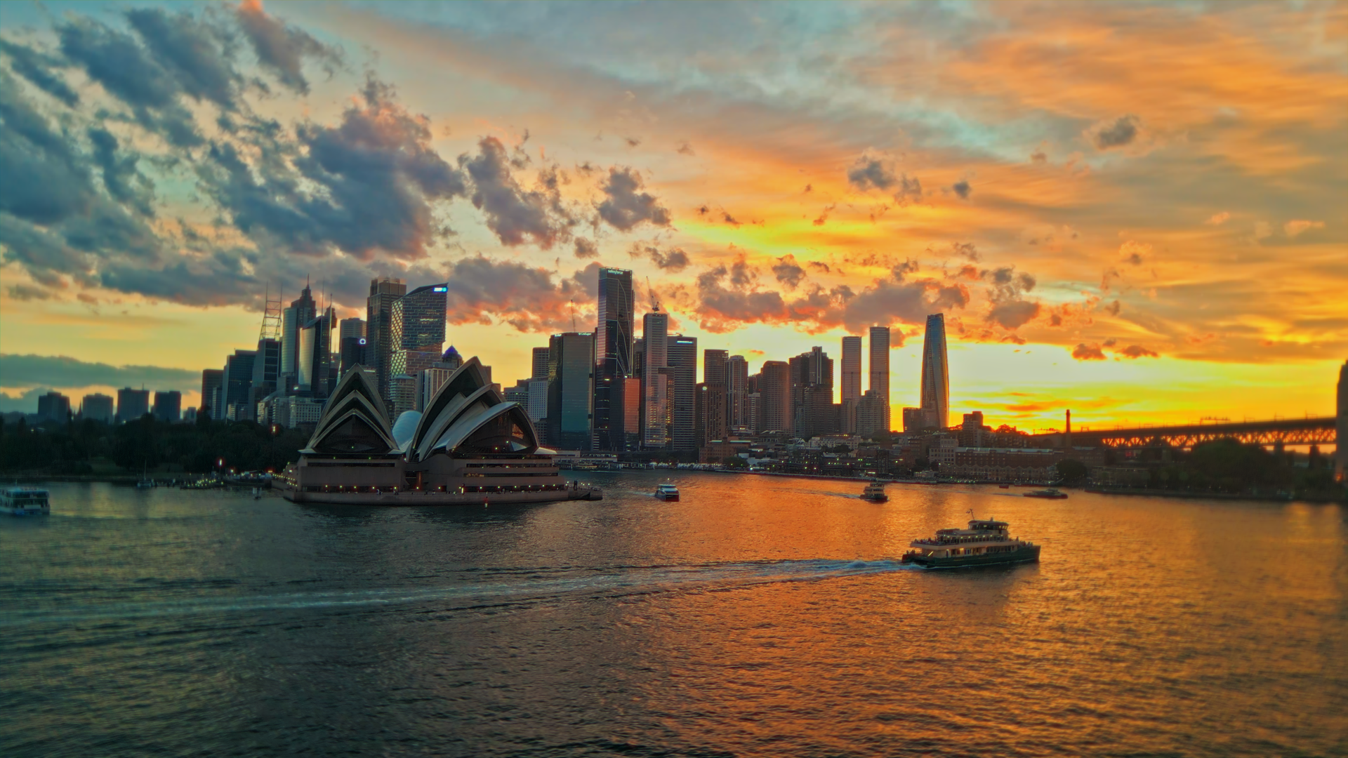 Uno skyline urbano con edifici moderni e il famoso Opera House di Sydney al tramonto, con barche sulla baia e nuvole nel cielo.