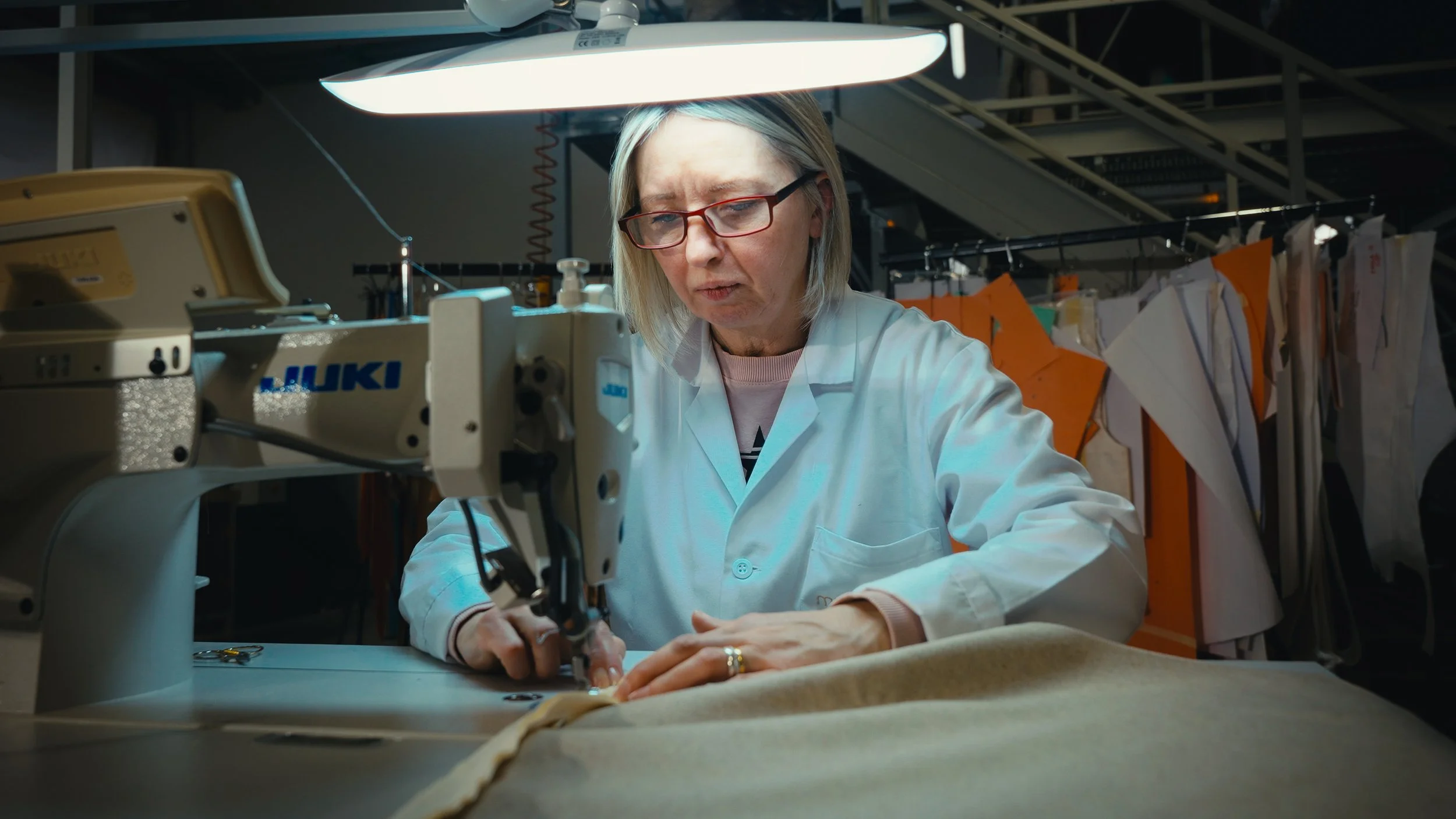 Una donna anziana con occhiali rossi che lavora su una macchina da cucire in un laboratorio di sartoria, indossando una giacca bianca.