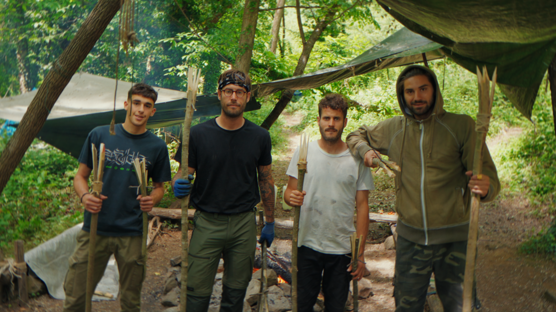 Quattro giovani uomini in un campeggio immerso nella natura, che tengono bastoni e posizionano un fuoco acceso.