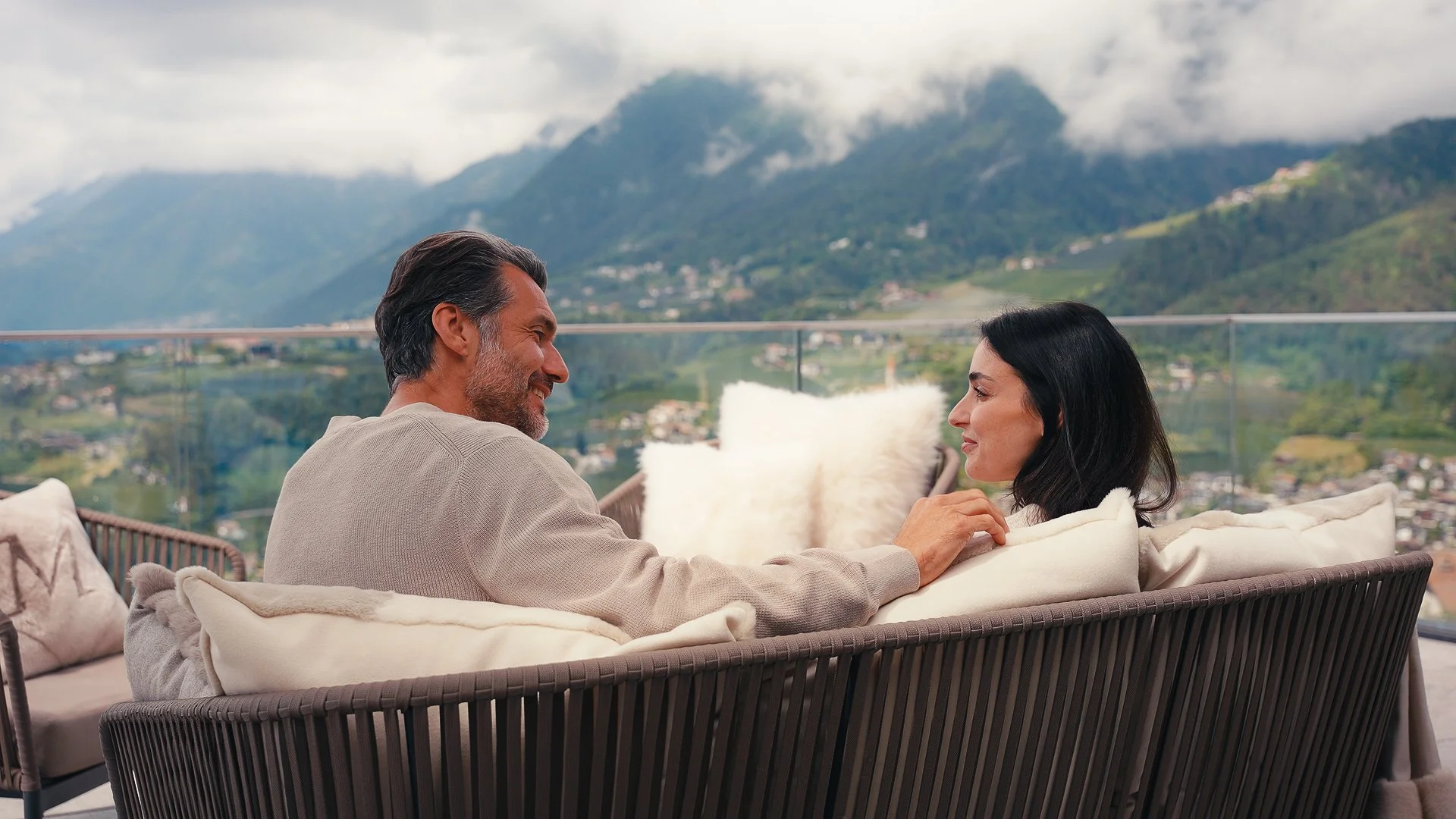 Una coppia di persone che si guarda sorridendo su un divano su un terrazzo con vista sulle montagne.