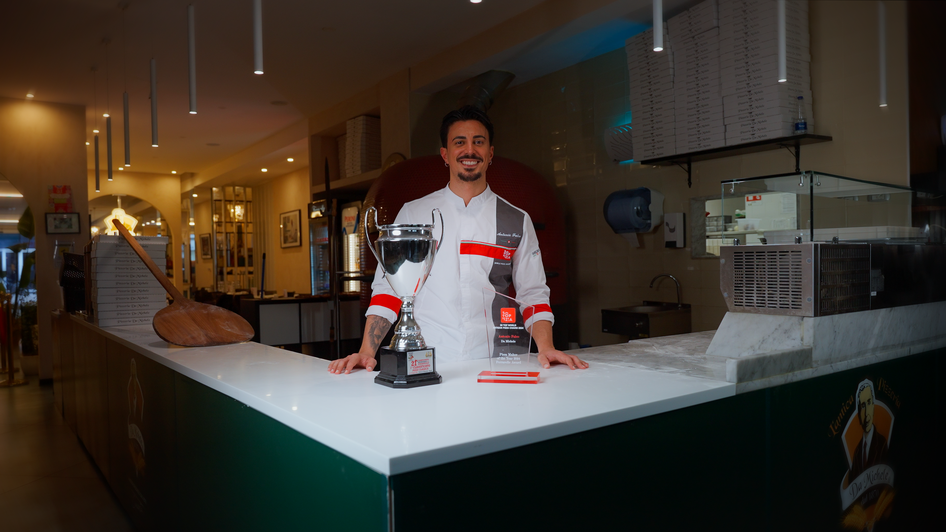 Cuoco sorridente dietro il bancone di un ristorante con un grande trofeo di cucina e una mini statuetta. Sul banco ci sono anche una spatola di legno, una scatola di pizza e una macchina per granite.