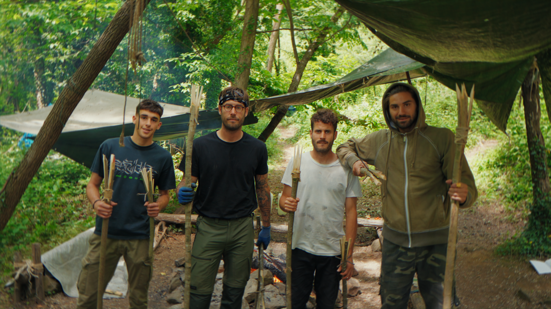 Quattro uomini in un campeggio nel bosco, con tende e legna, che tengono bastoni.