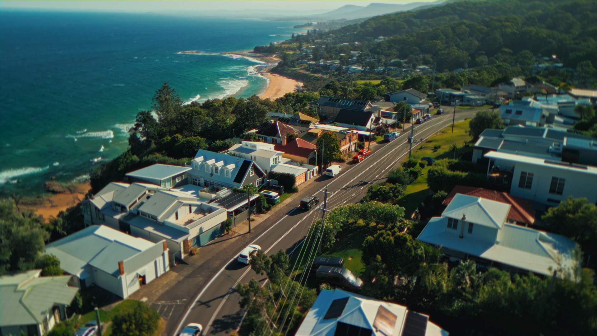 Veduta aerea di una costa con case, strada e mare, con colline verdi sullo sfondo.
