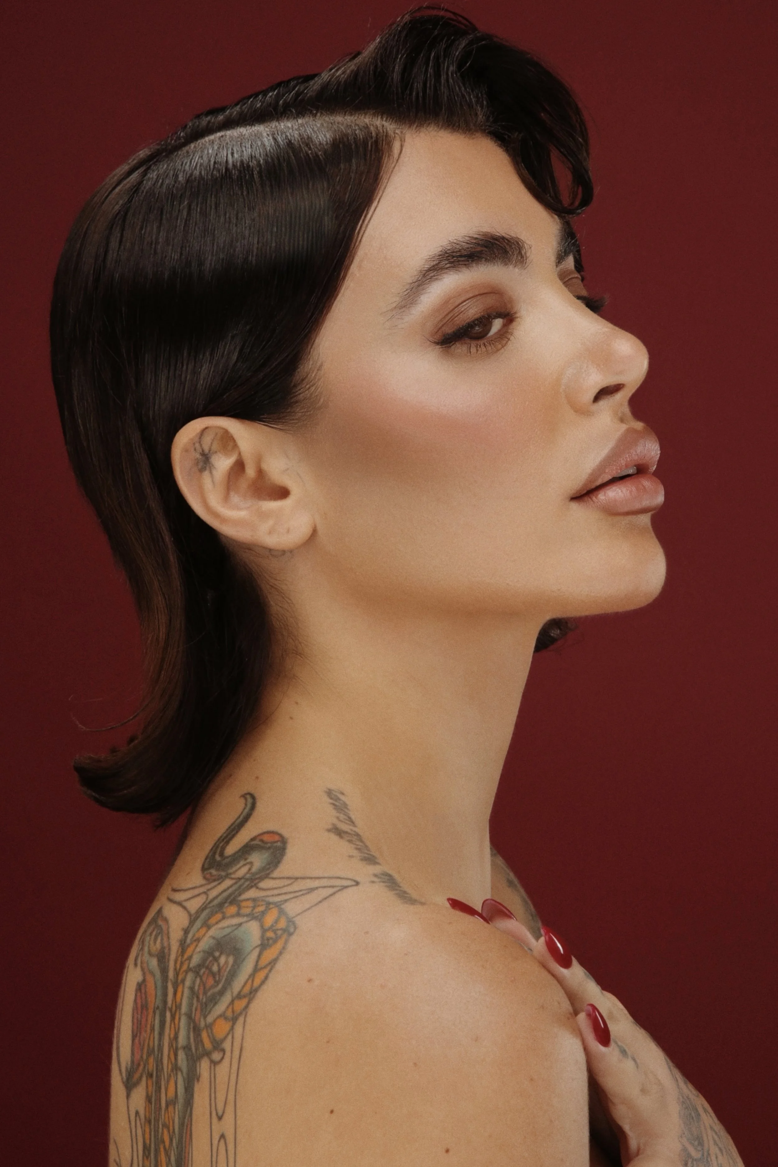 Ritratto di una donna con capelli corti, trucco elegante e tatuaggi sulla spalla e sul collo, sfondo rosso.