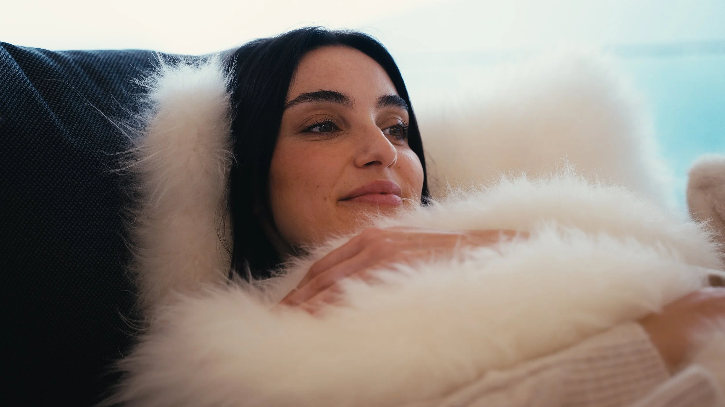 Una donna con orecchie di pelliccia nera e vestiti di pelliccia bianca è sdraiata su un divano, con uno sfondo sfocato di un ambiente luminato.