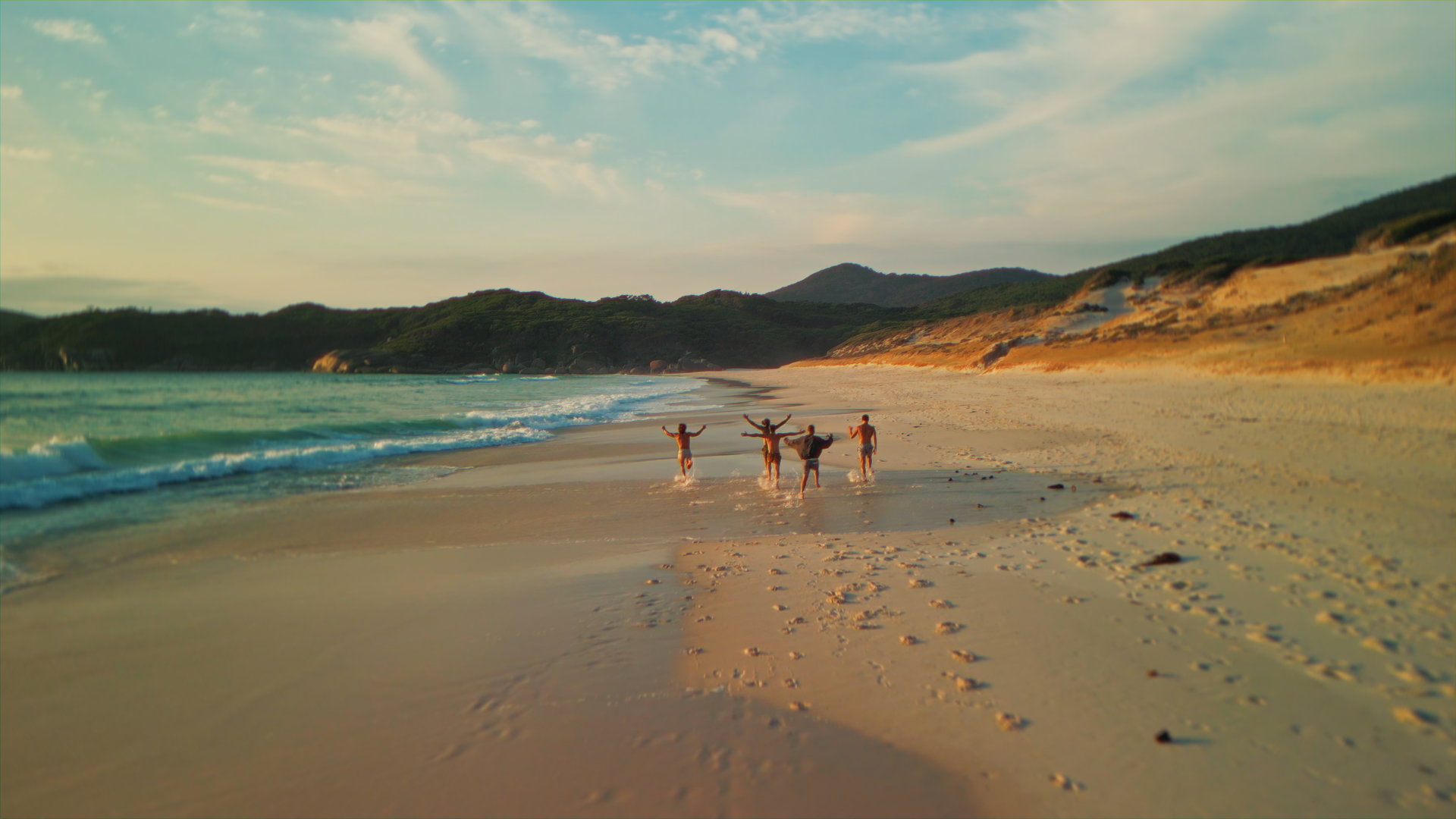 Gruppo di cinque persone che camminano sulla spiaggia al tramonto, con onde che si infrangono e colline verdi sullo sfondo.