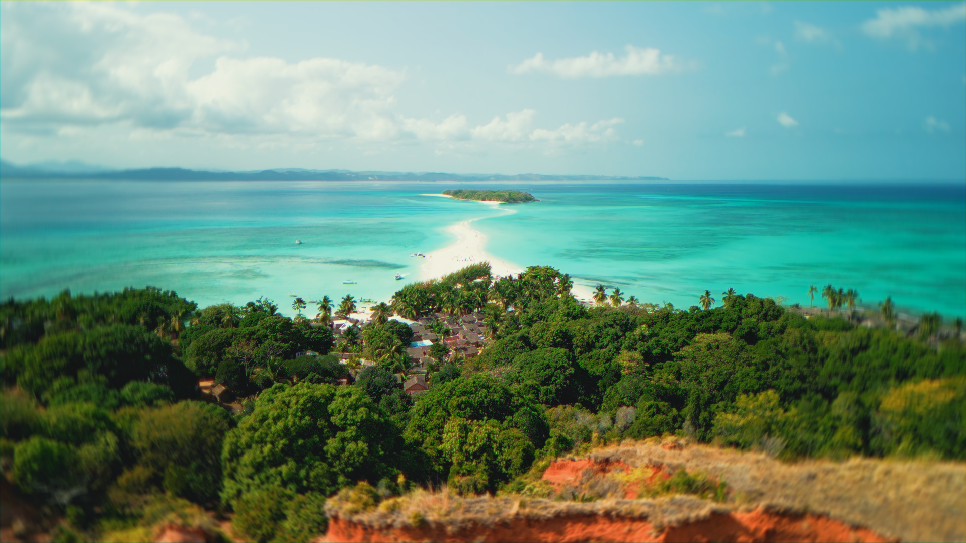 Veduta aerea di un'isola tropicale con mare turchese, spiaggia bianca e vegetazione verde, con alcune barche in acqua.