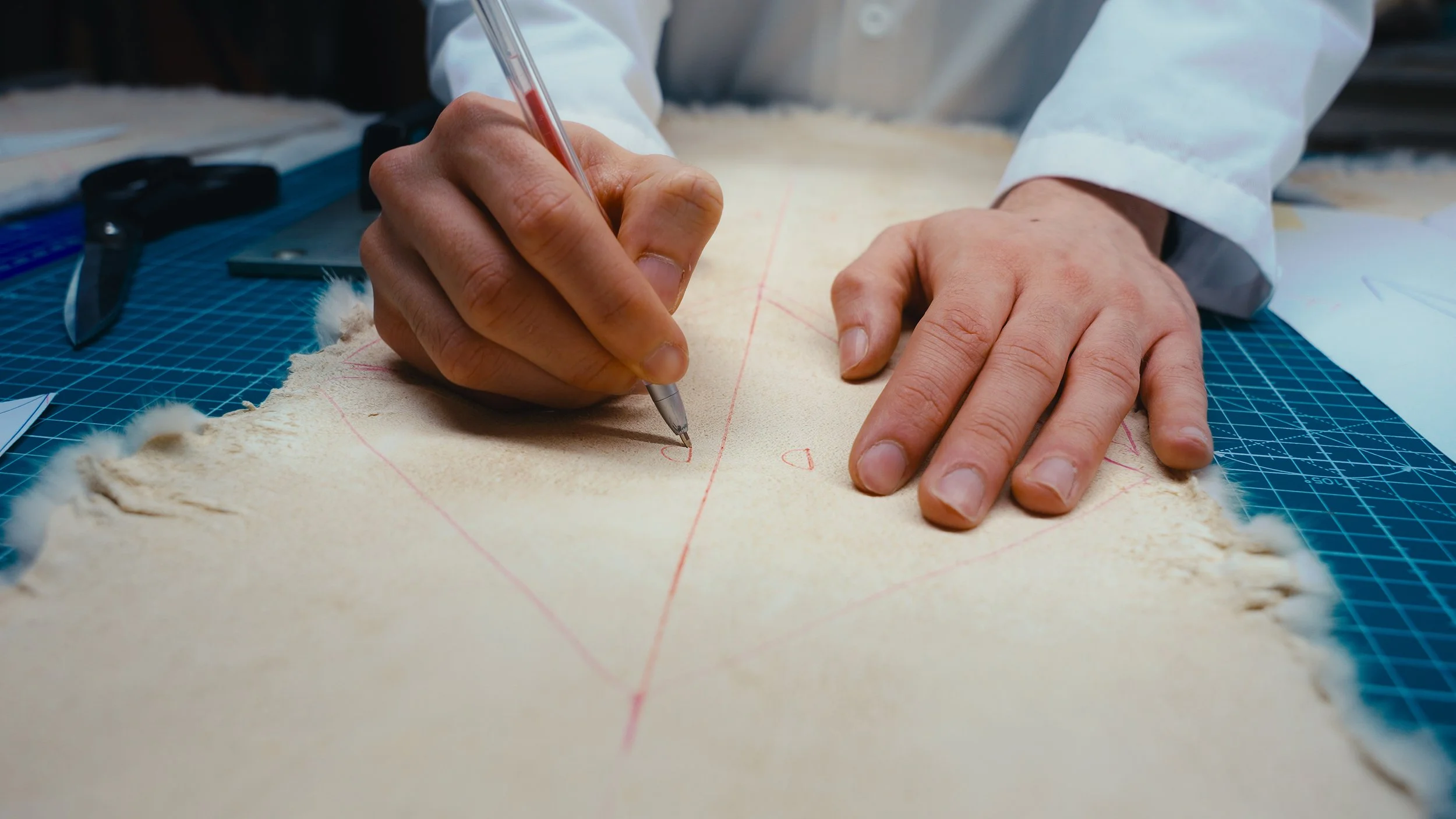 Una persona che traccia linee rosse su un tessuto di pelle con un penna a inchiostro rosso, sul tavolo ci sono forbici e un righello.