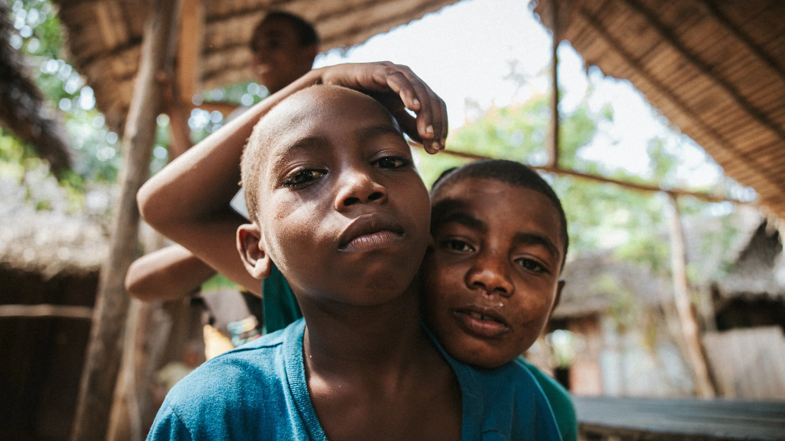 Due giovani bambini con pelle scura, uno con capelli corti e l'altro con una ciocca di capelli, guardano seriamente verso la fotocamera in un ambiente diurno sotto un tetto di paglia, con uno sfondo di case di legno e albero.