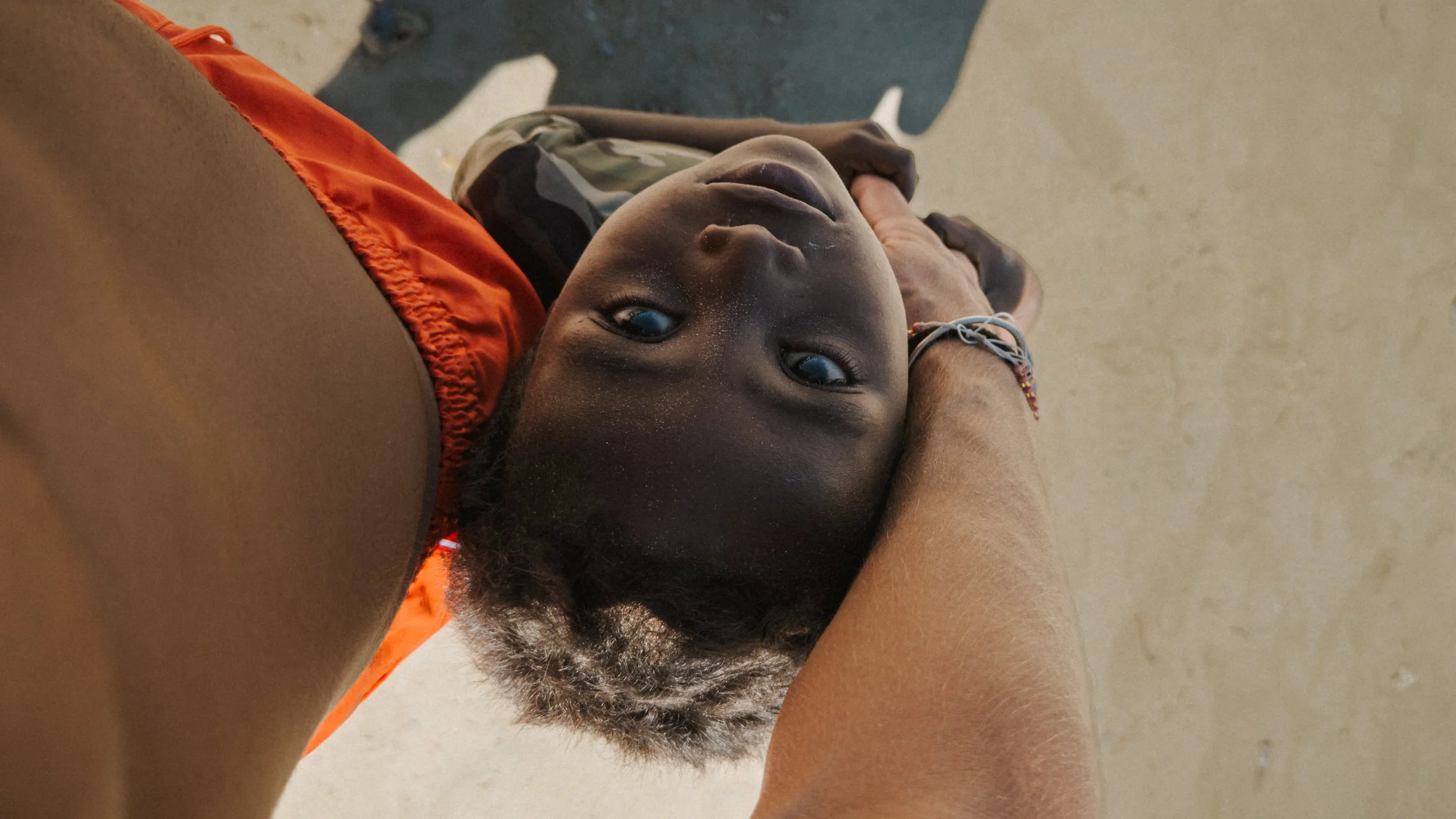 Ragazzo con pelle scura sdraiato sulla spalla di una persona, guarda verso la fotocamera, con sfondo neutro.