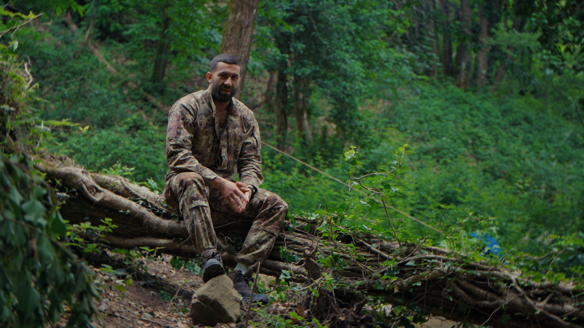 Uomo in uniforme mimetica seduto su un albero caduto nel bosco verde.