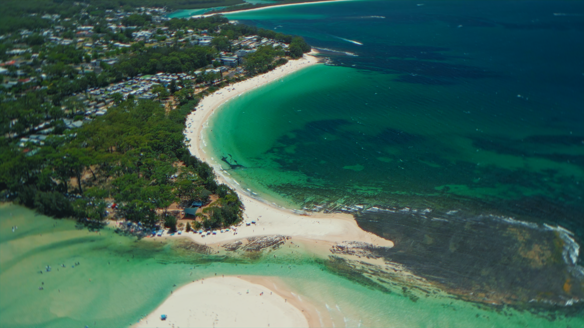 Immagine aerea di una spiaggia con sabbia bianca, acque turchesi e una zona di mare più scura, circondata da vegetazione e insediamenti umani.