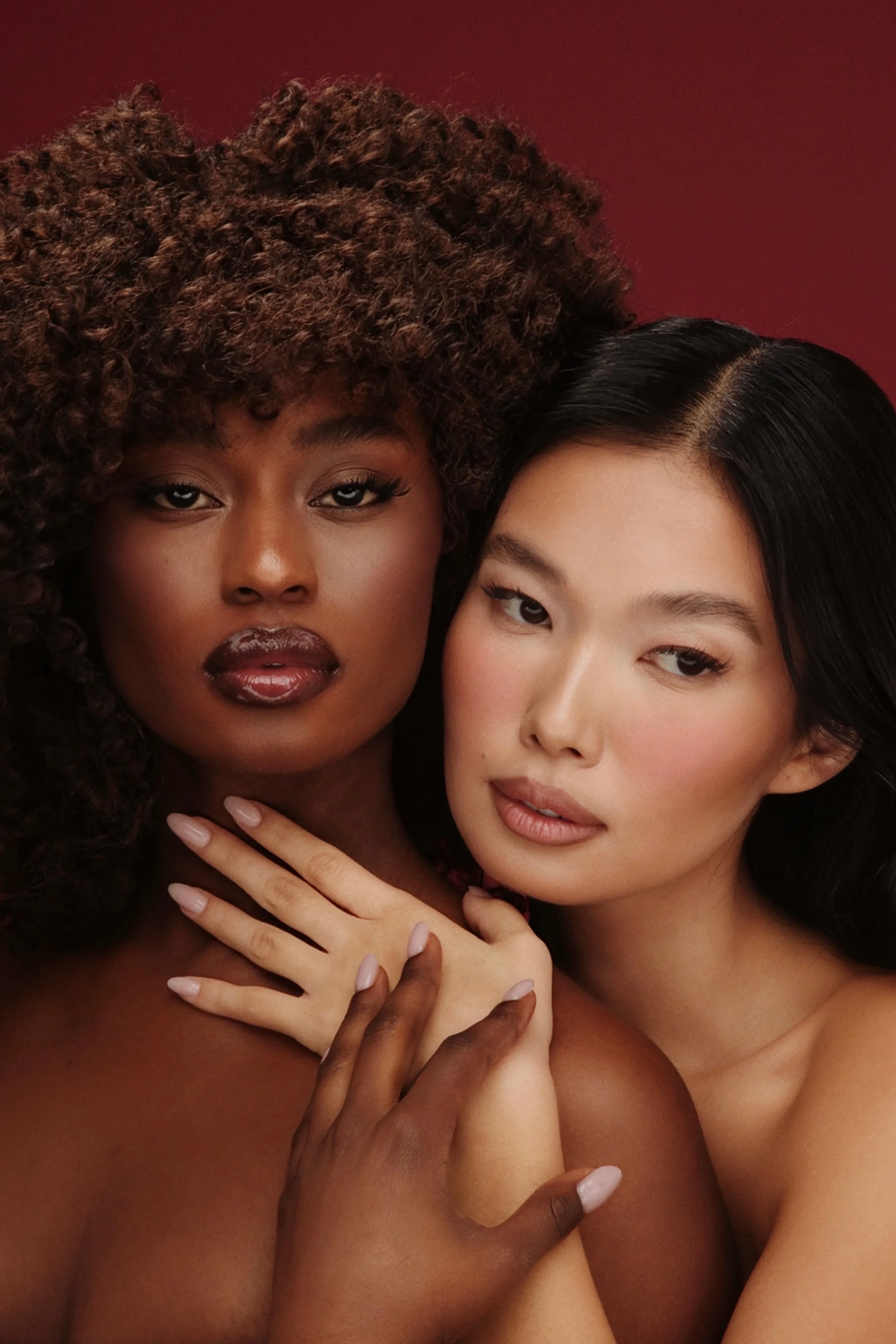 Due donne senza vestiti si abbracciano, una con capelli ricci e scuri e l'altra con capelli lisci e neri, con sfondo rosso.