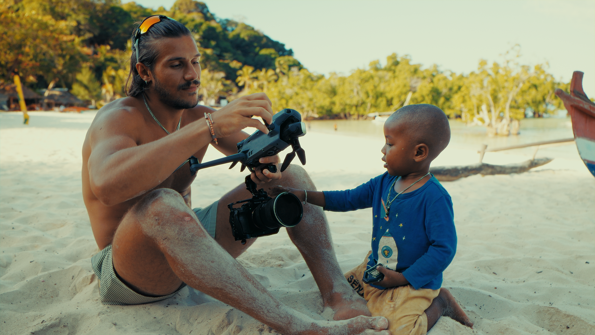 Un uomo e un bambino sono seduti sulla spiaggia, l'uomo tiene un drone e il bambino tocca il drone, sullo sfondo ci sono alberi e barche.