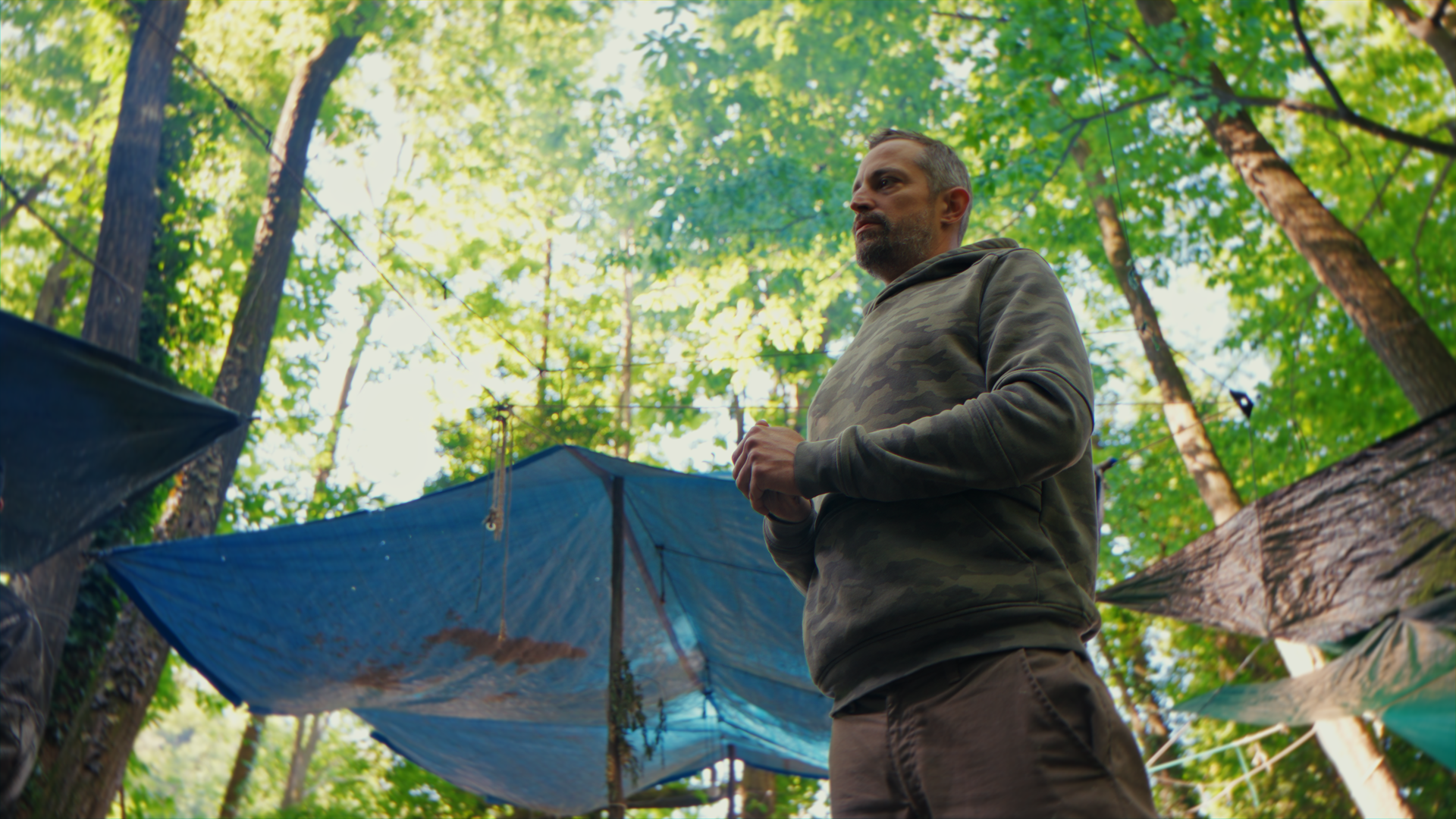 Uomo con cappuccio grigio in un bosco, circondato da tende blu e altri rifugi, in un ambiente naturalistico