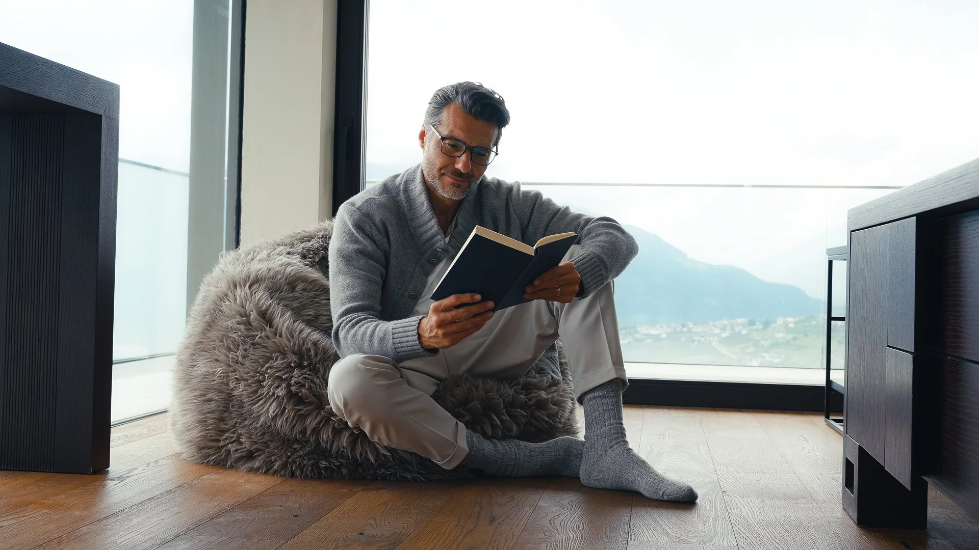 Un uomo anziano con capelli grigi, occhiali, maglione grigio e pantaloni chiari si siede sul pavimento di legno con una coperta di pelliccia soffice, leggendo un libro vicino a una grande finestra con vista sulle montagne e sul paesaggio.