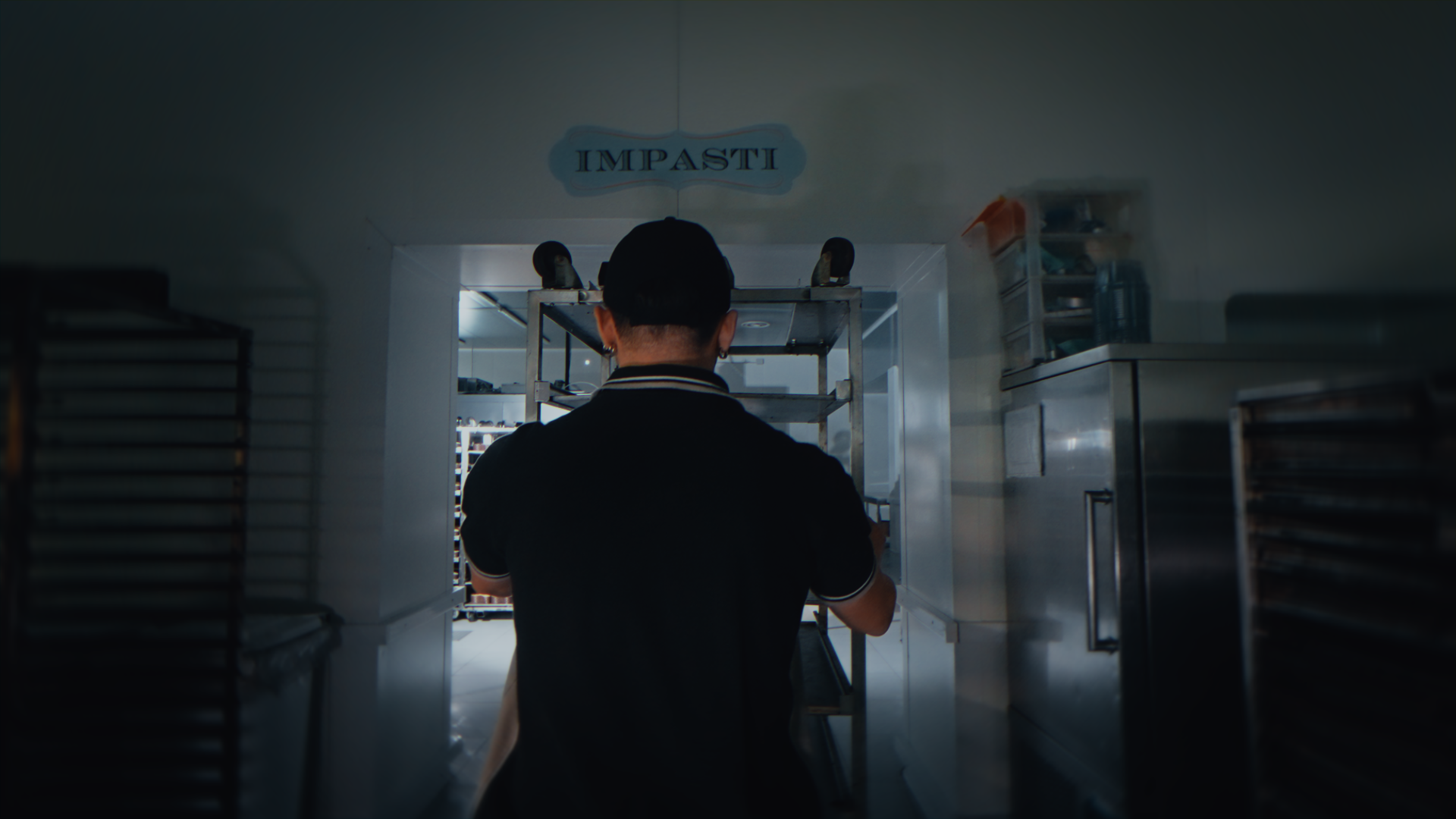 Una persona vista di schiena che si trova in una cucina, sotto un cartello con la scritta 'IMPASTI'.