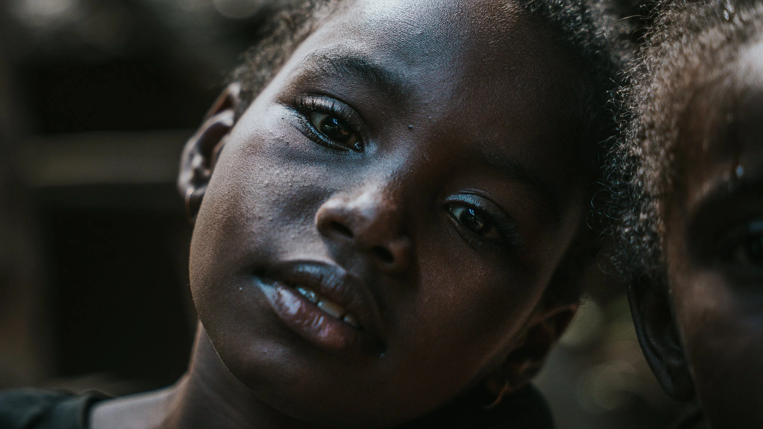 Ragazza con pelle scura, occhi marroni e capelli ricci, guarda verso la fotocamera.
