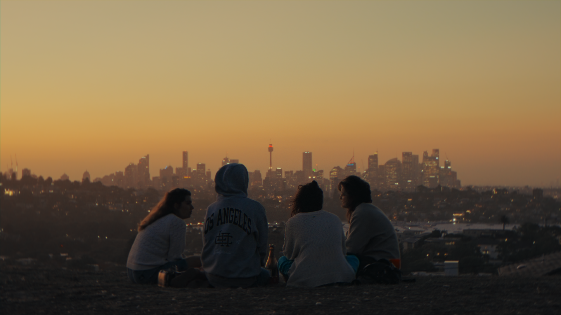 Quattro persone sedute sul terreno durante il tramonto con la skyline di Los Angeles sullo sfondo.