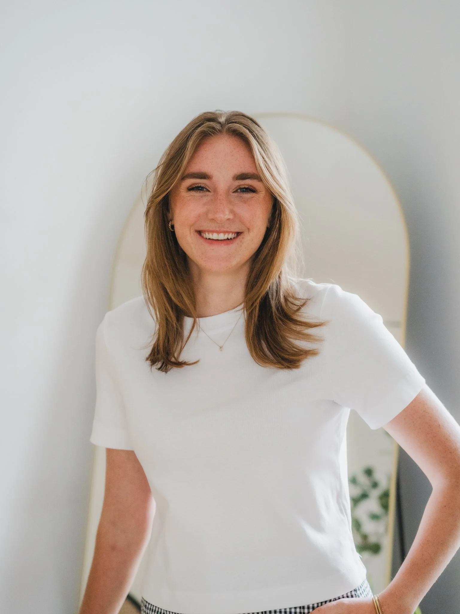 A young woman with shoulder-length light brown hair, smiling, wearing a white T-shirt and gold jewelry, standing in front of a mirror with a gray wall background.
