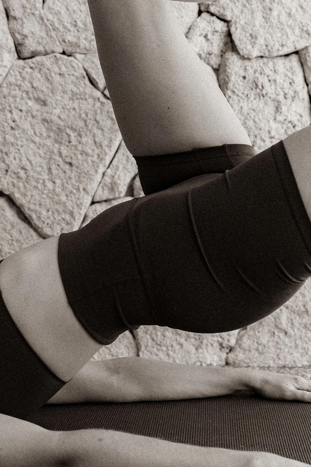 Close-up of a person practicing yoga or stretching, with one leg extended and resting on the floor against a stone wall, wearing black athletic clothing.