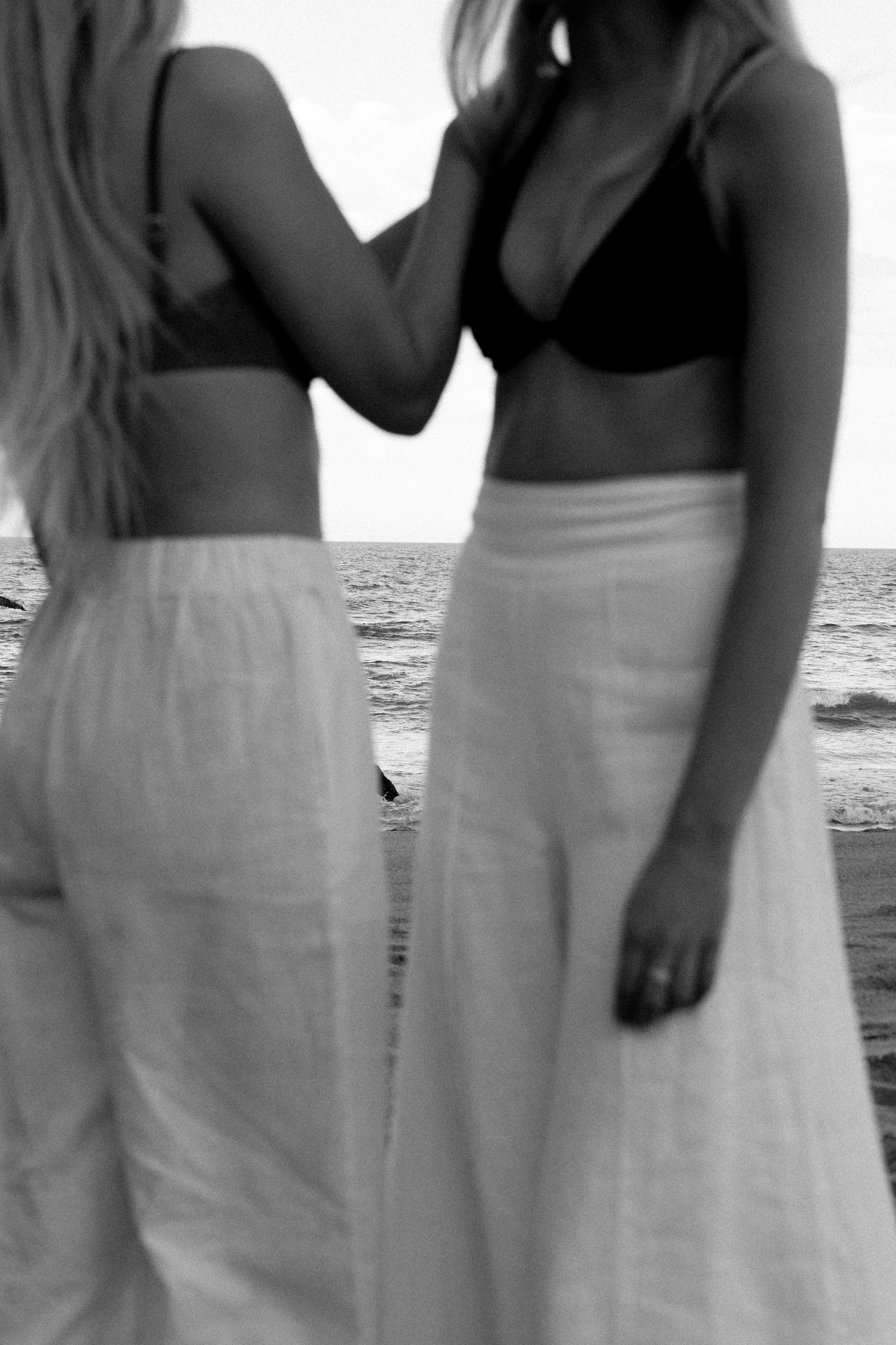 Two women in swimsuits standing on a beach near the ocean, holding hands and embracing.