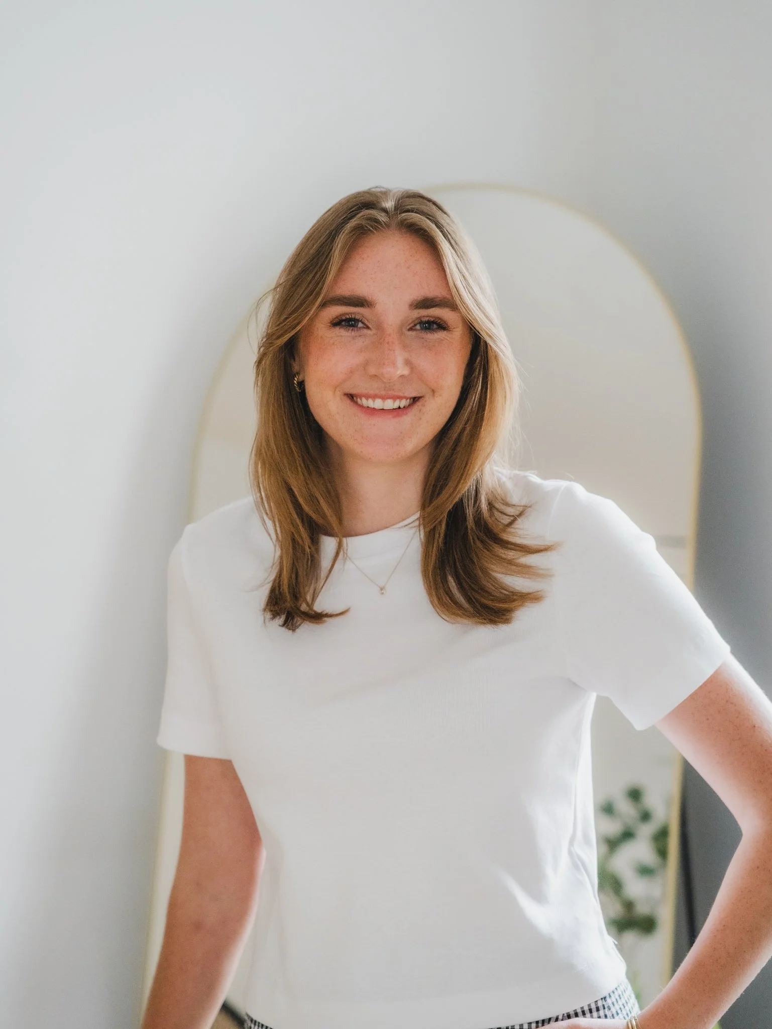 A smiling young woman with shoulder-length brown hair, wearing a white t-shirt, standing indoors with a mirror and some plants in the background.