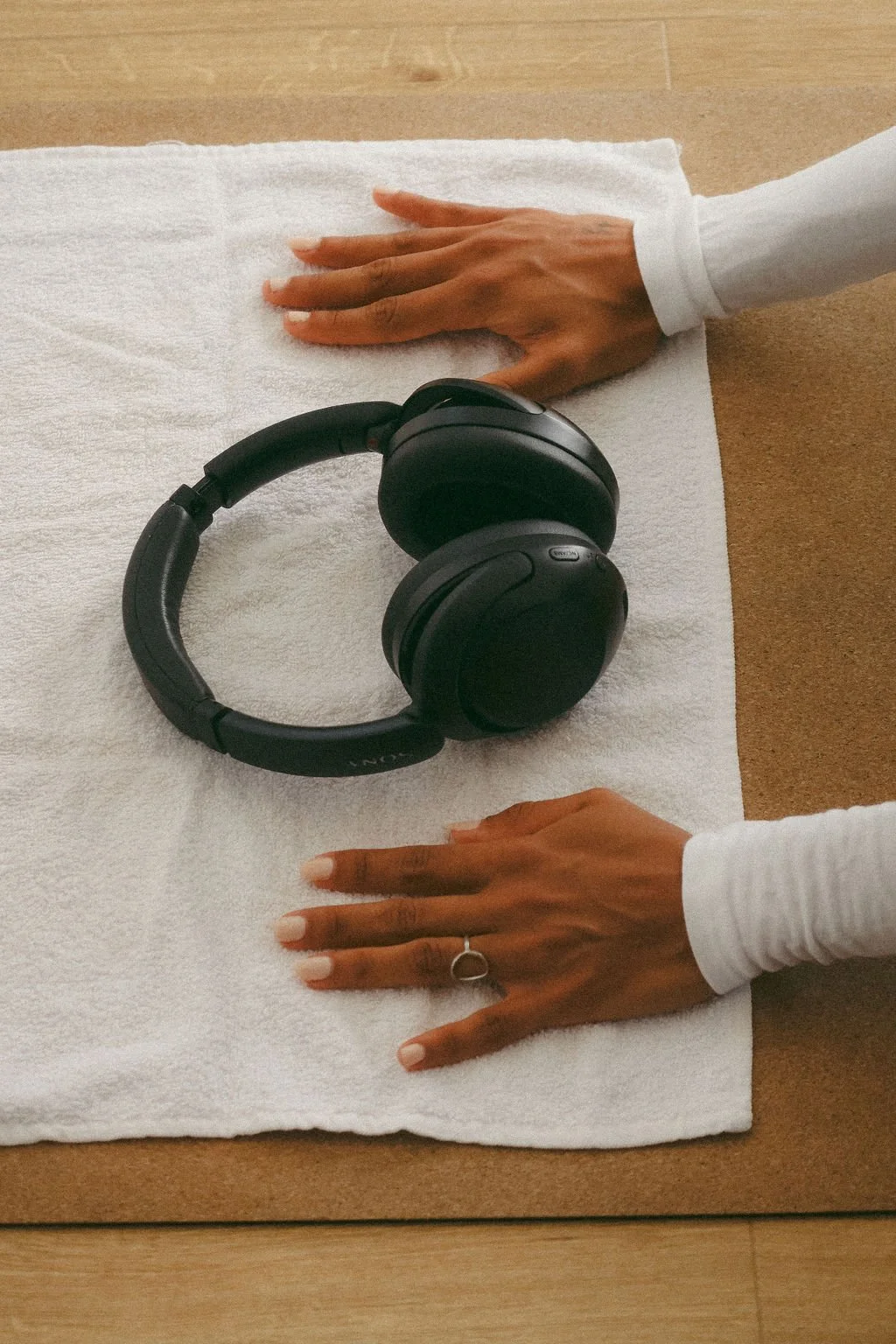 Person with medium skin tone and long nails, wearing a white long-sleeve shirt and a ring on the left hand, laying hands flat on a white towel on a wooden surface with black over-ear headphones in between.