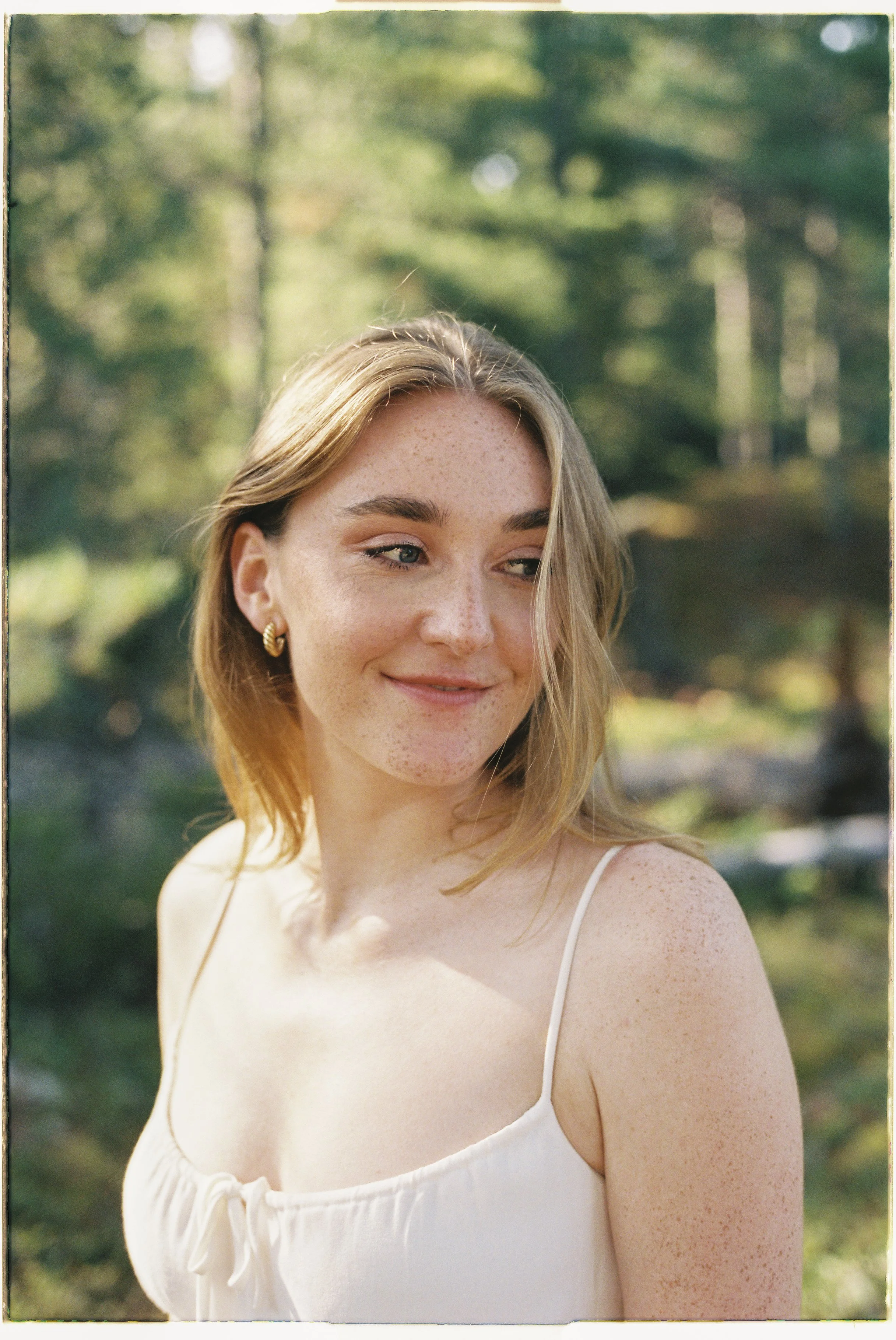 A young woman with light brown hair, freckles, and gold hoop earrings, smiling slightly while looking to the side. She is outdoors in a natural setting with trees in the background, wearing a white spaghetti strap top.