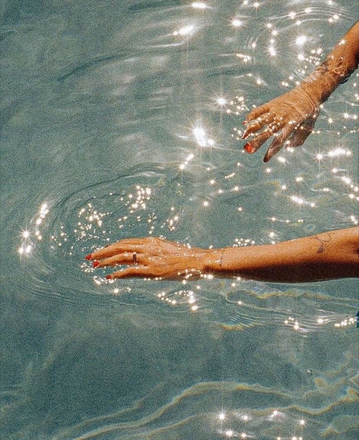 Two hands, one above the water and one below, reaching towards each other in a swimming pool with sunlight reflecting off the surface.