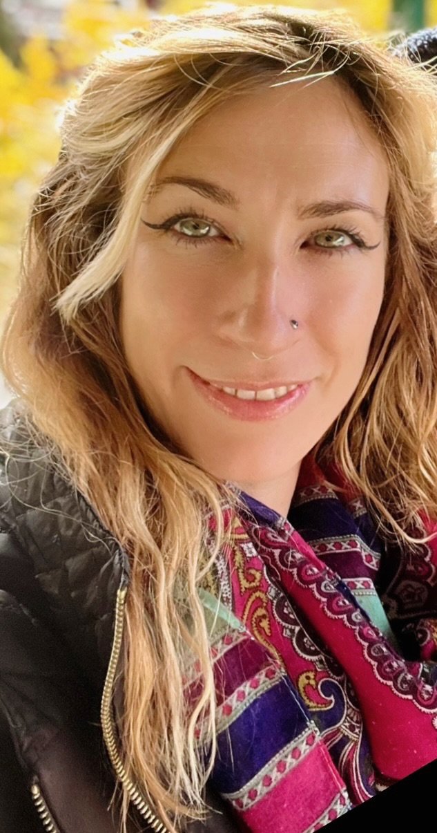 A young woman with blonde hair, green eyes, a nose piercing, wearing eye makeup, and a colorful scarf, smiling at the camera.