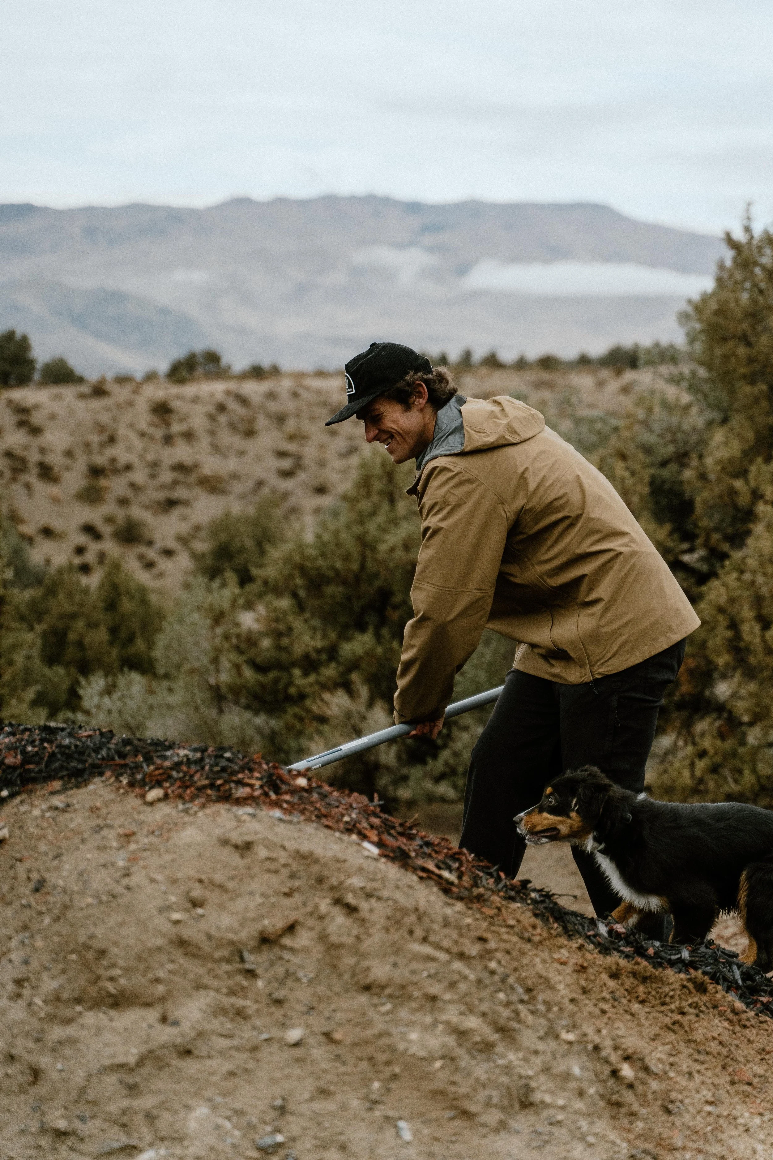 A man wearing a beige jacket and black cap, smiling while using a shovel on a dirt mound in a desert landscape with mountains and sparse vegetation, accompanied by a black and tan dog.