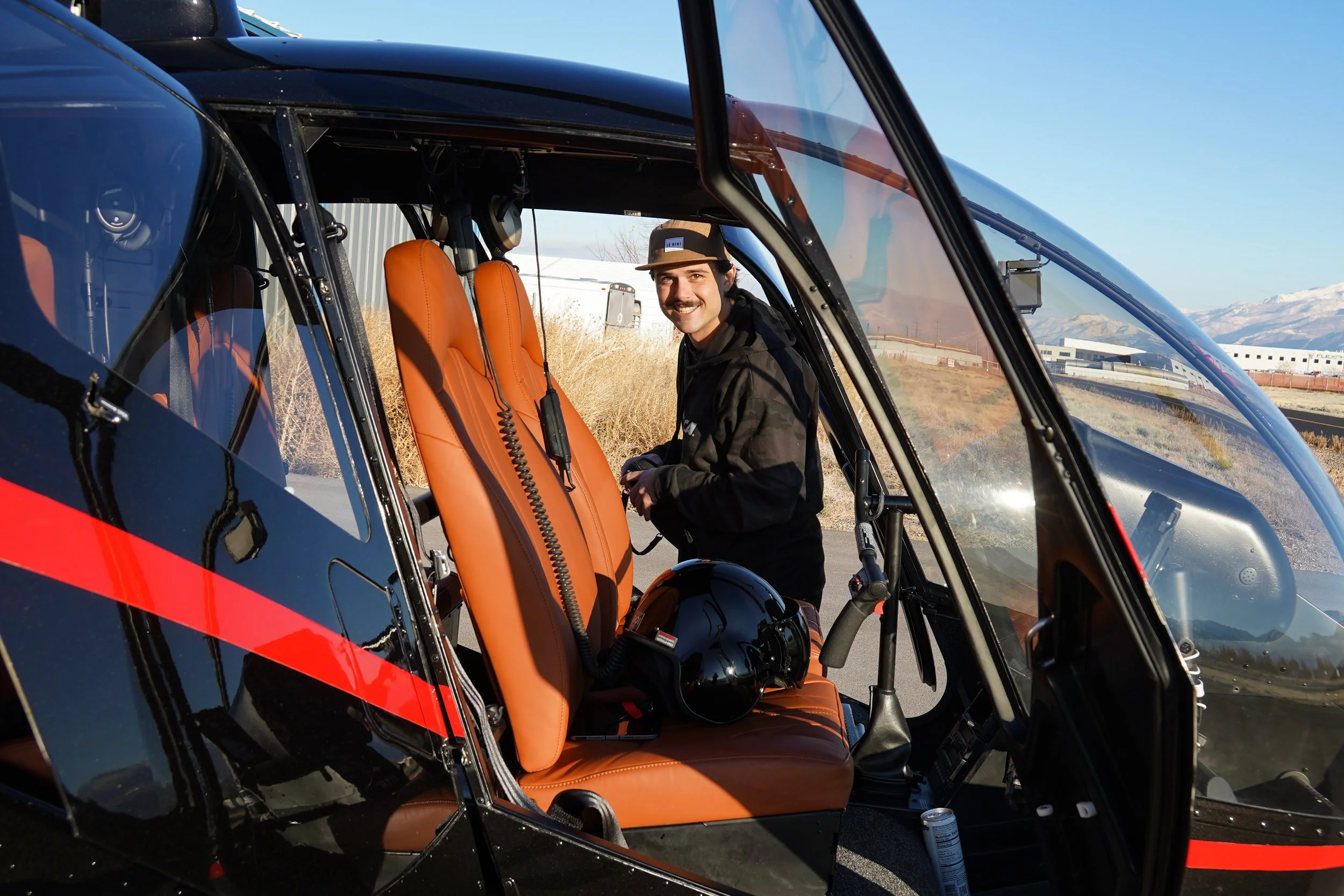 A man with a mustache and wearing a black jacket and a gray hat inside a helicopter with brown leather seats, smiling at the camera.