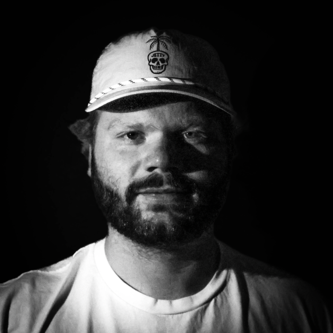 Black and white photo of a man with a beard wearing a cap with a skull and palm tree logo, and a plain T-shirt, against a dark background.