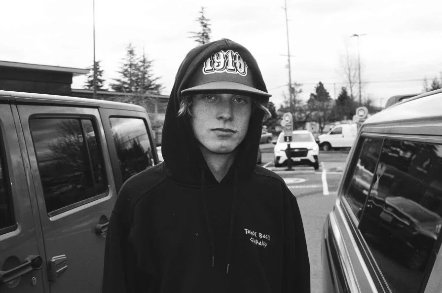 A young man with light skin and light-colored hair stands in a parking lot, wearing a hooded sweatshirt and a baseball cap, with a serious expression.
