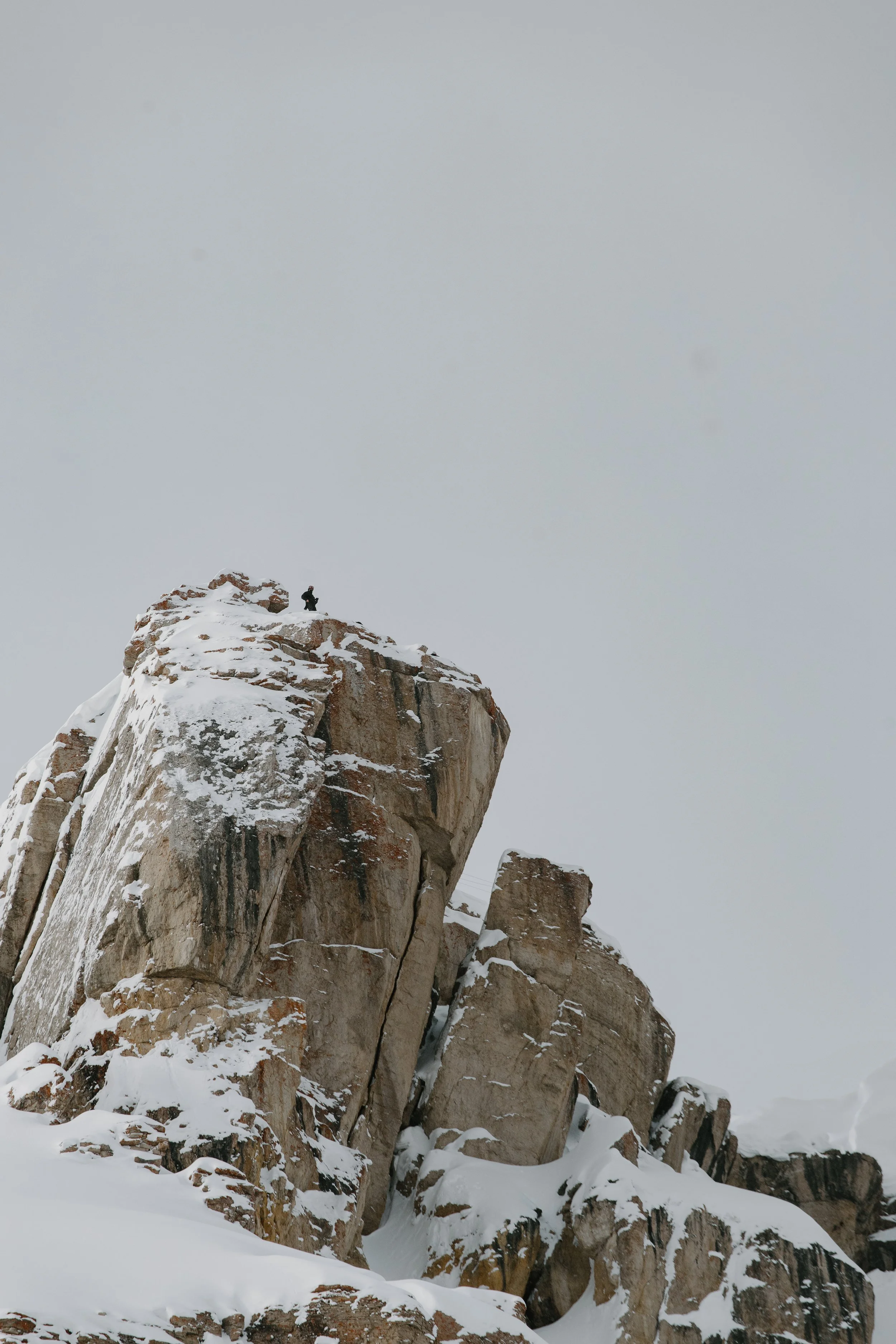 A person standing on top of a snow-covered rocky mountain peak against a cloudy sky.
