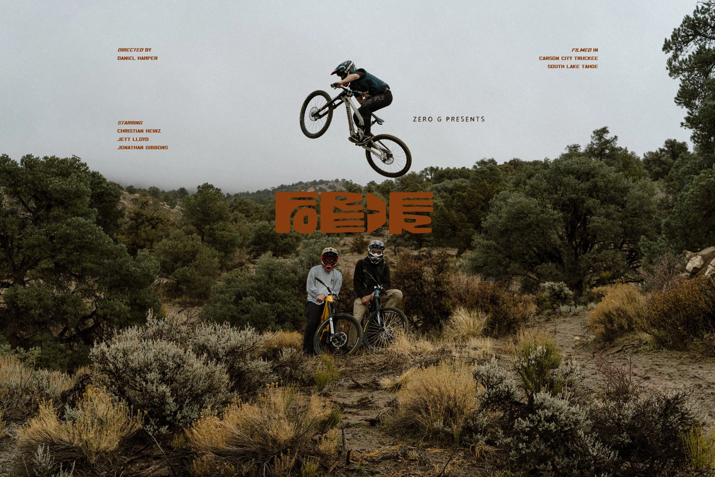 Two mountain bikers standing on a trail in a desert landscape with bushes and trees, one of them wearing a white jacket and the other a black jacket, while a third rider in mid-air performs a jump on a bicycle against a cloudy sky.