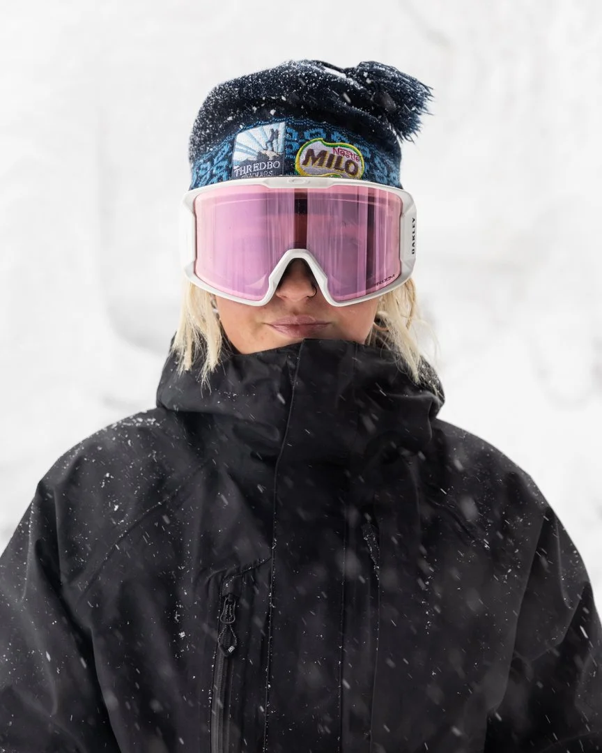 Person wearing ski goggles, a winter hat, and a black winter jacket, standing in a snowy environment.