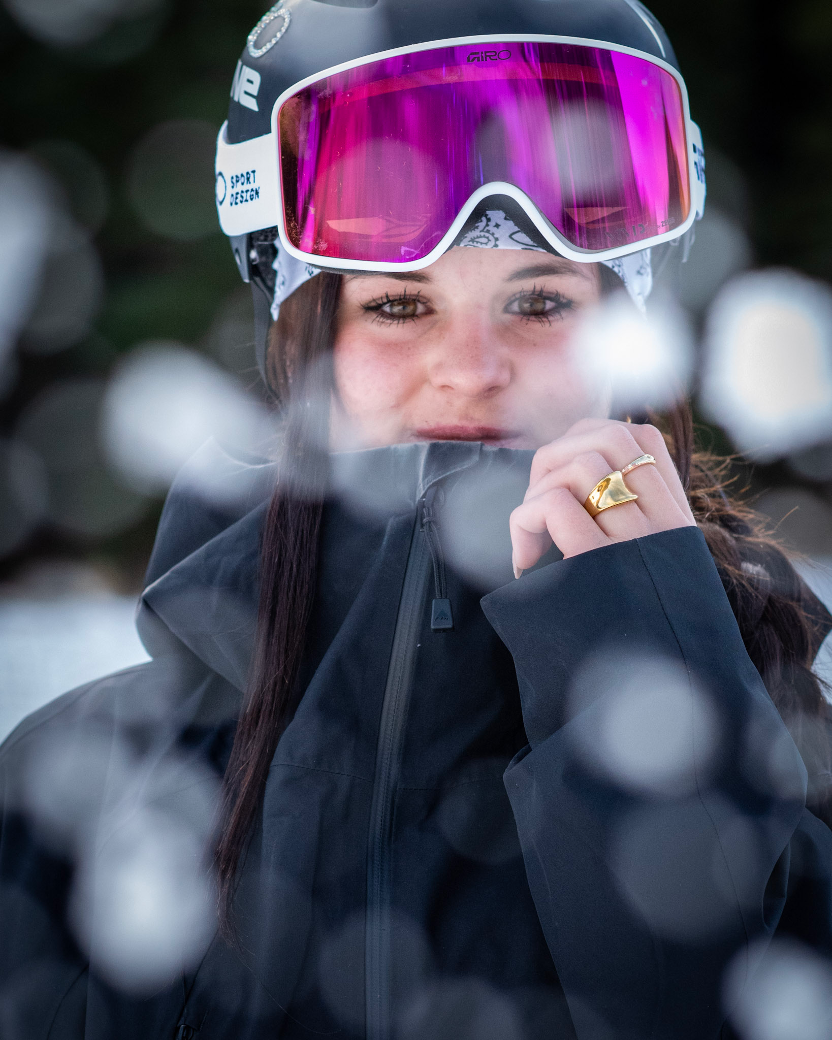A woman wearing a black jacket, a white bandana, and a ski helmet with pink-tinted goggles looks directly at the camera, holding her jacket out with her hand, which has a gold ring on her finger, with blurred background.